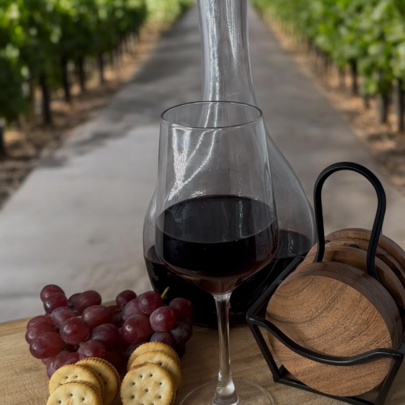 Glass of red wine and decanter with grapes and crackers on a wooden board, vineyard in the background.