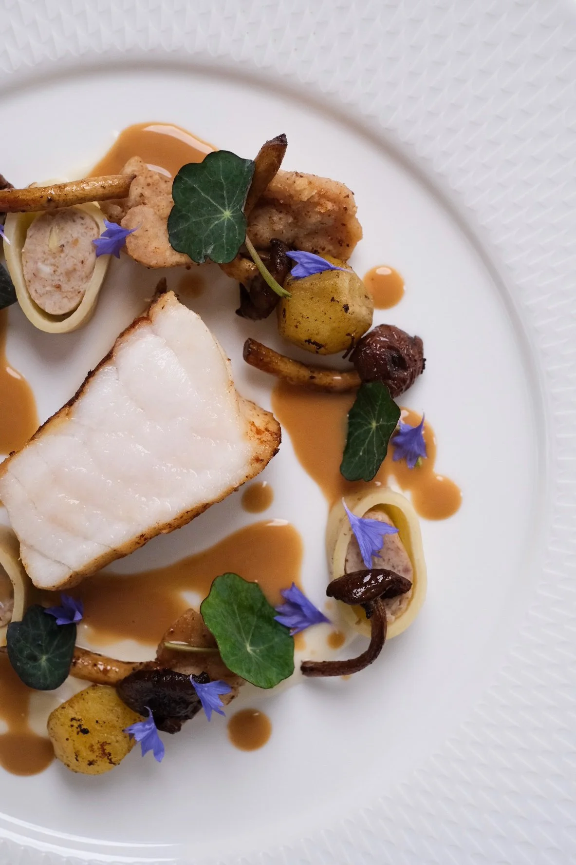 A plated gourmet dish with a piece of white fish, garnished with edible blue flowers, green leaves, and sprigs of mushrooms, with a light brown sauce drizzled around.