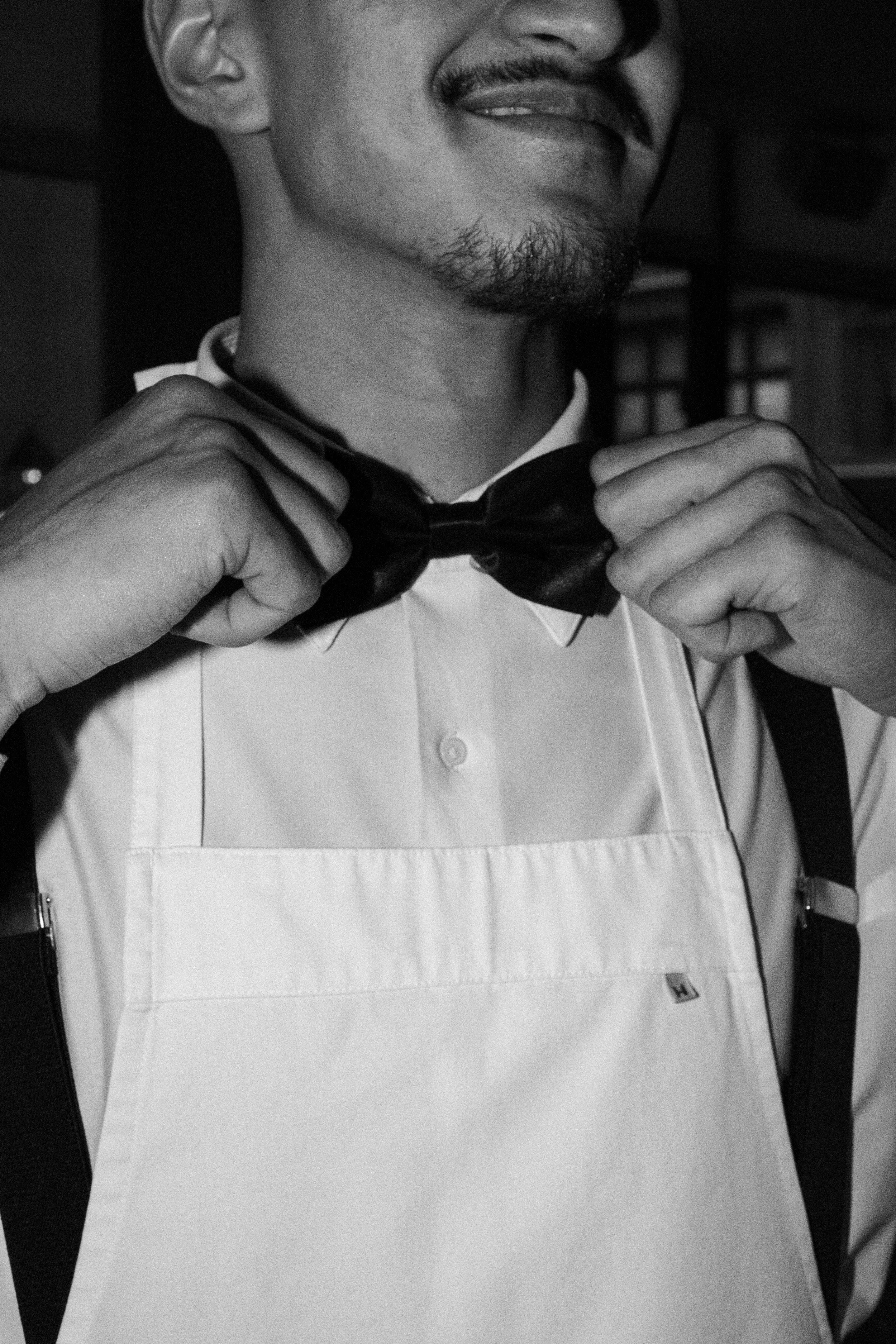 A man adjusting his bow tie, wearing a white shirt, black suspenders, and an apron. His face is partially visible, showcasing a mustache and goatee, in a black and white photograph.