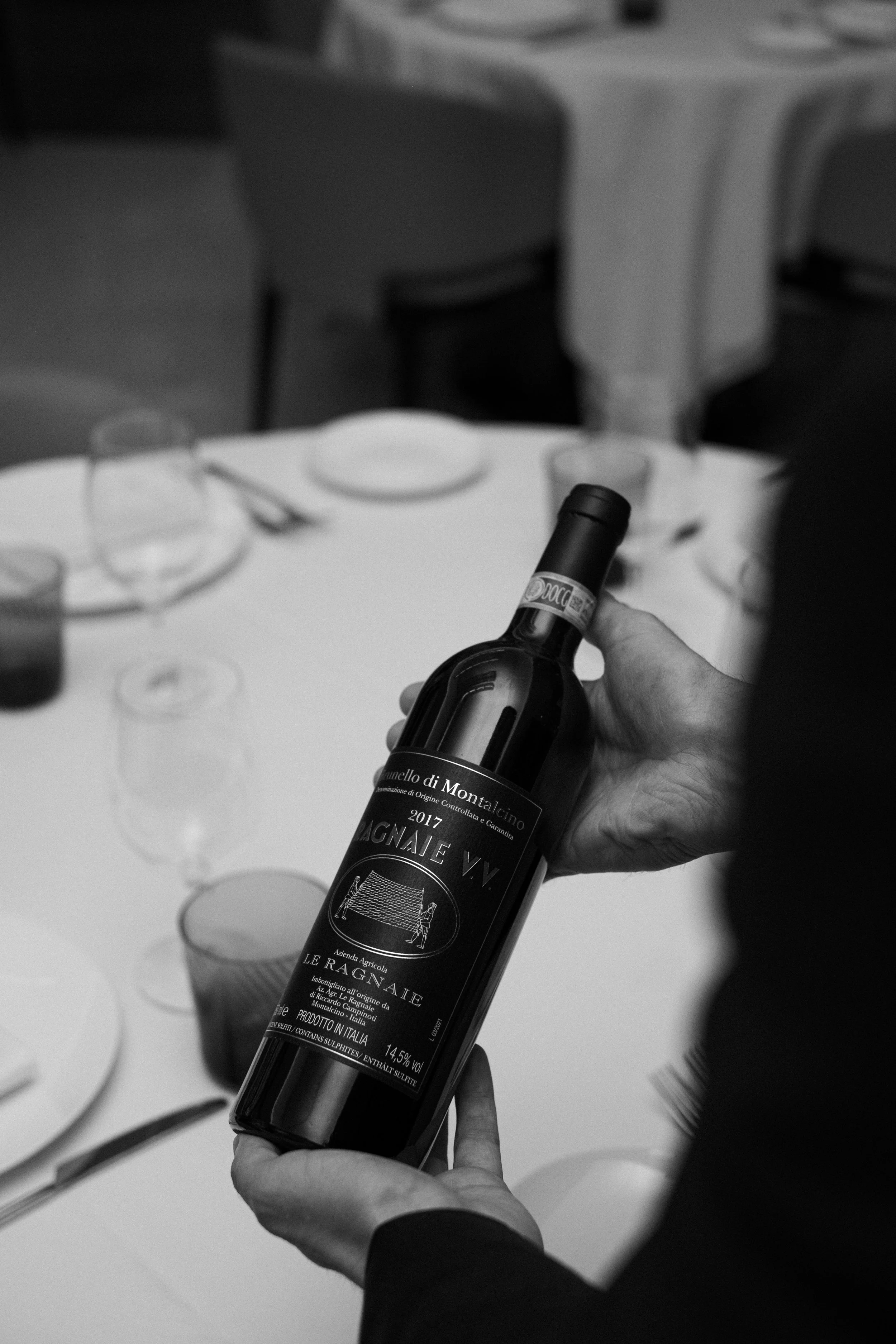 A person holding a bottle of Italian red wine at a dining table set for a meal.