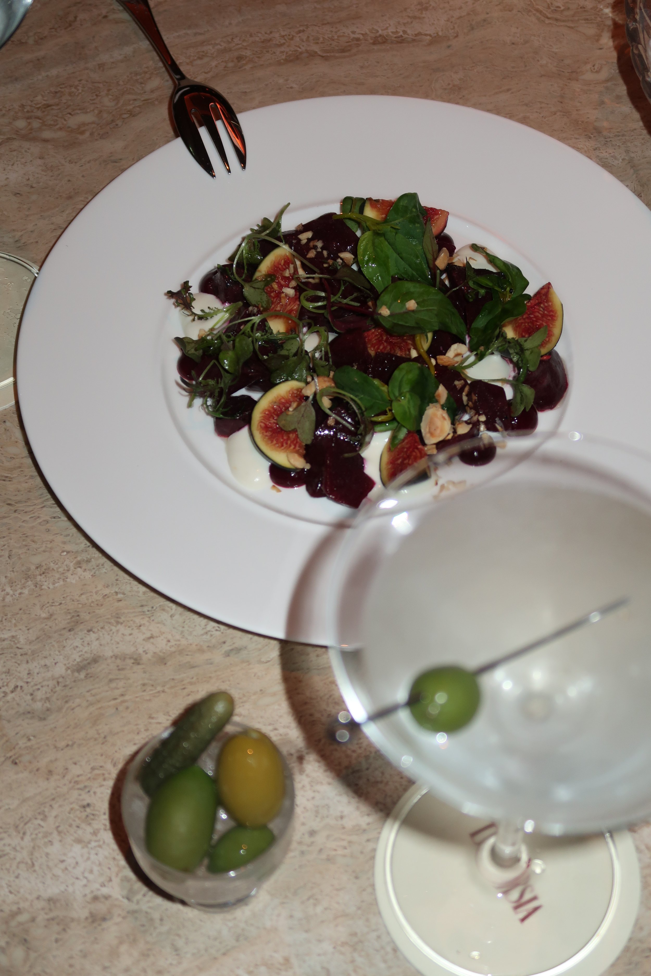 A white plate with a salad made of greens, figs, and nuts, topped with a balsamic glaze. A fork is placed on the top left of the plate. In the foreground, there is a cocktail glass with a green olive and a small glass of mixed olives, on a beige table.