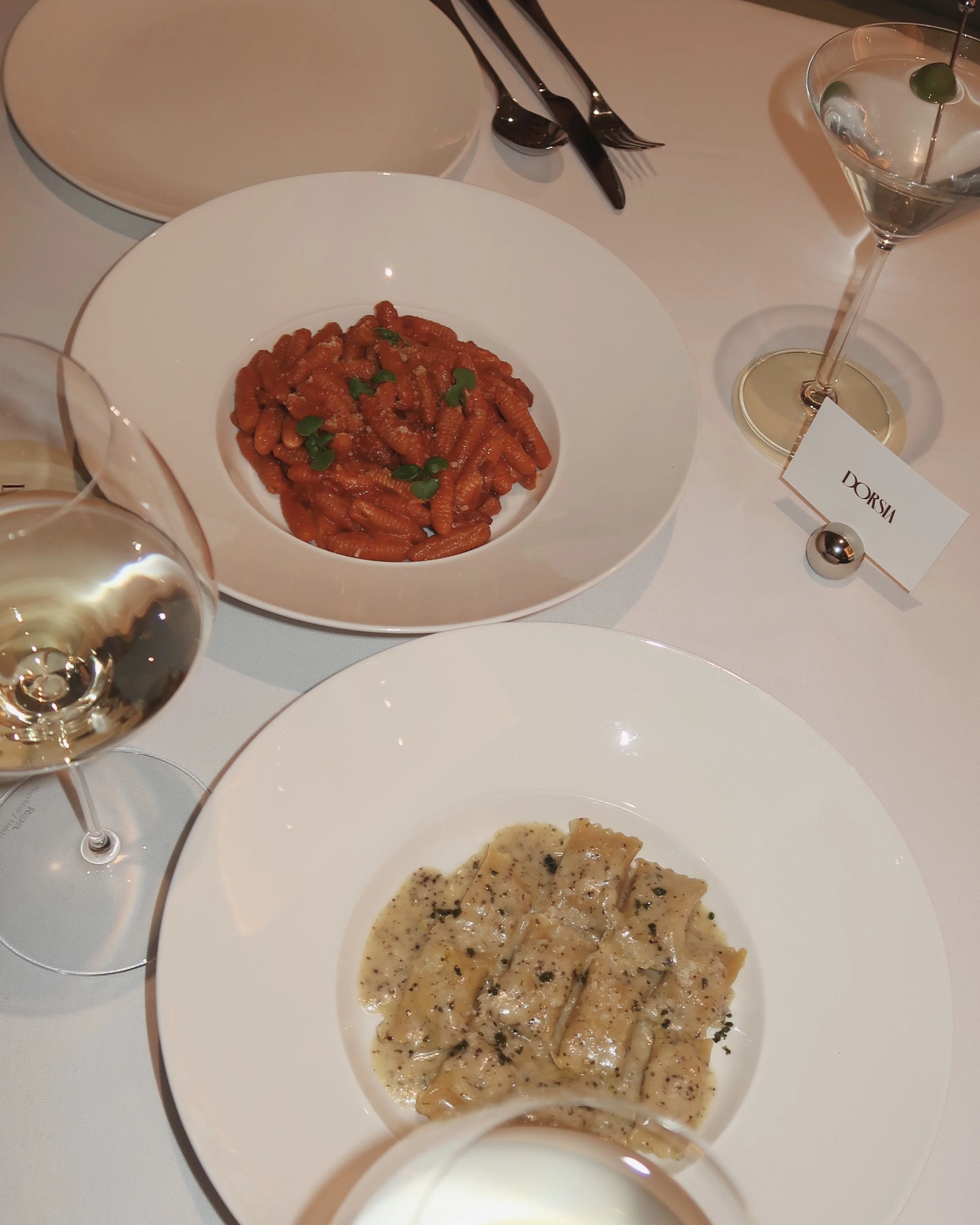 Plate of cooked pasta with sauce and green garnish, and a bowl of pasta with white sauce and herbs on a white tablecloth, with glasses of white wine and an empty plate in the background.