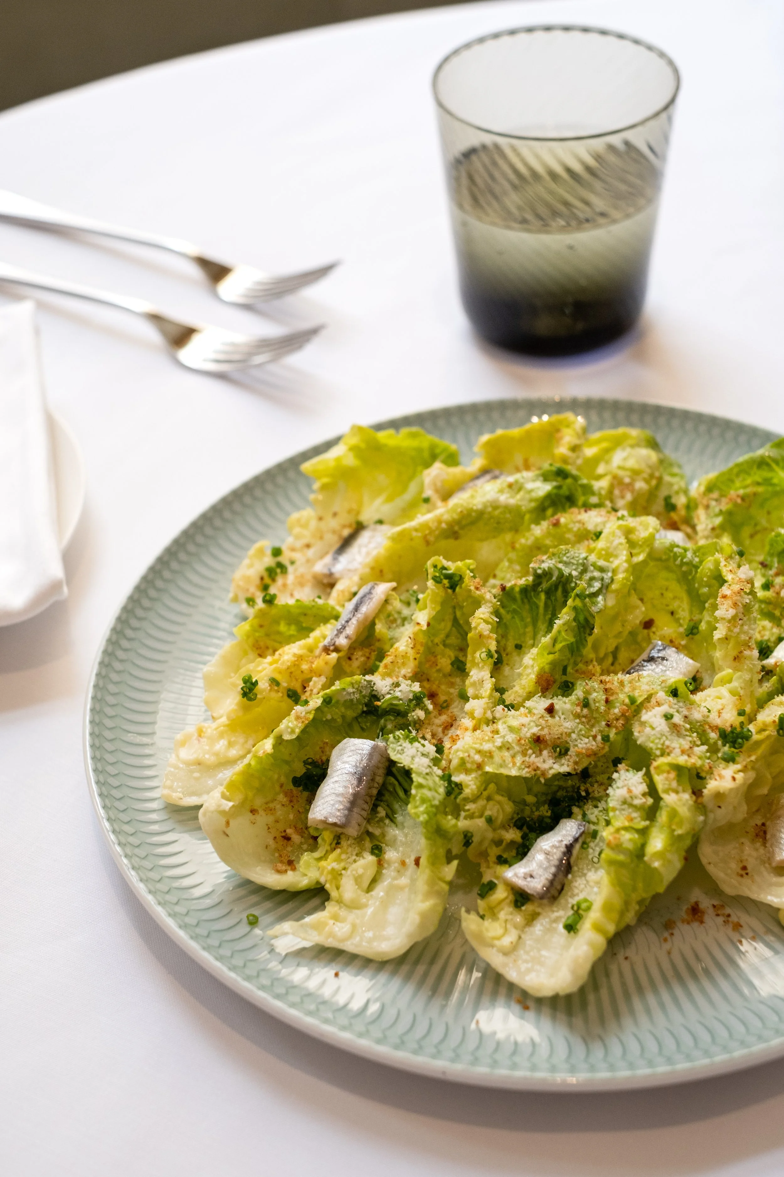 Caesar salad with lettuce, anchovies, and grated cheese on a green-rimmed plate, with a glass of dark soda and utensils on a white tablecloth.