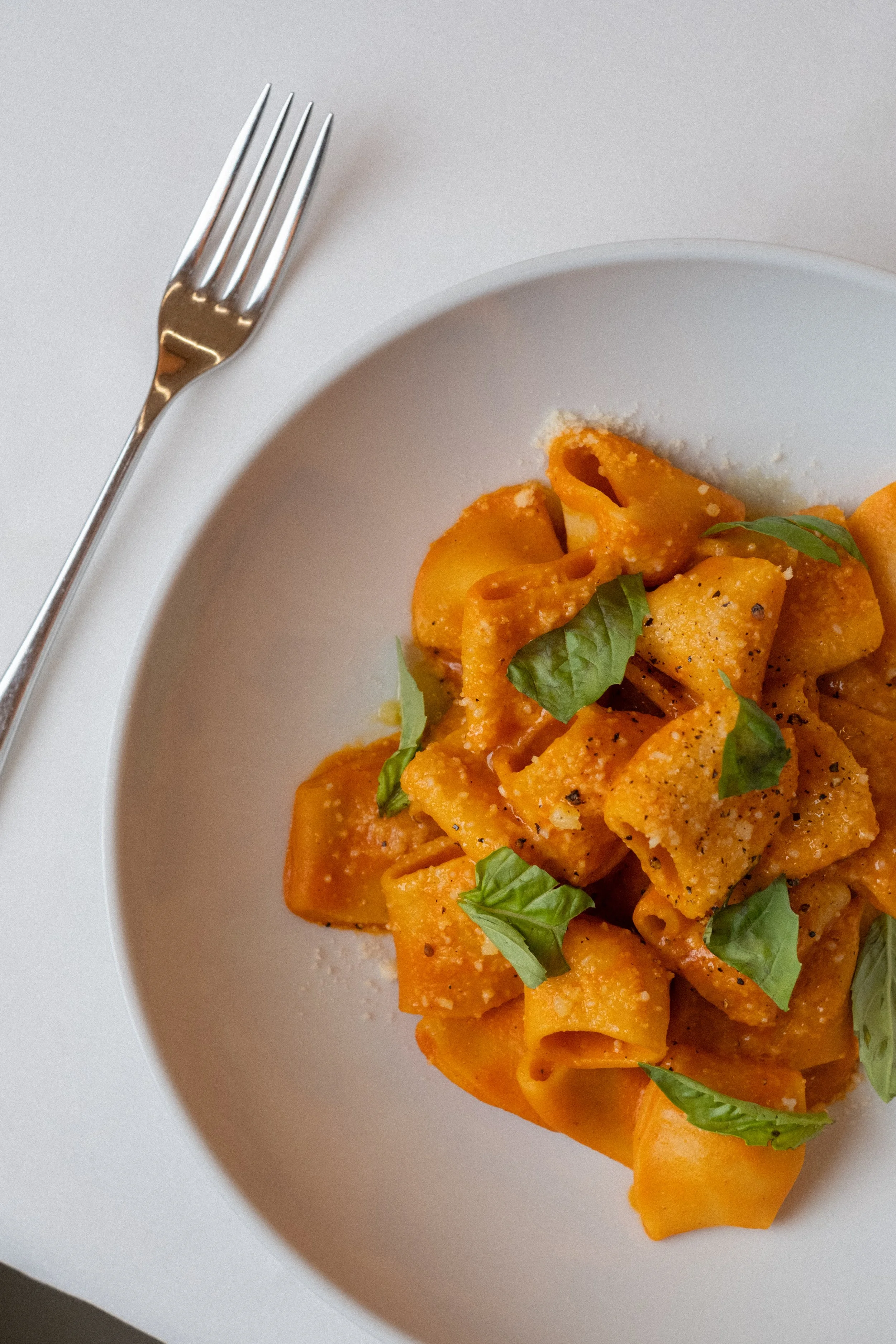 Plate of pasta with tomato sauce, garnished with fresh basil leaves, on a white table with a silver fork.