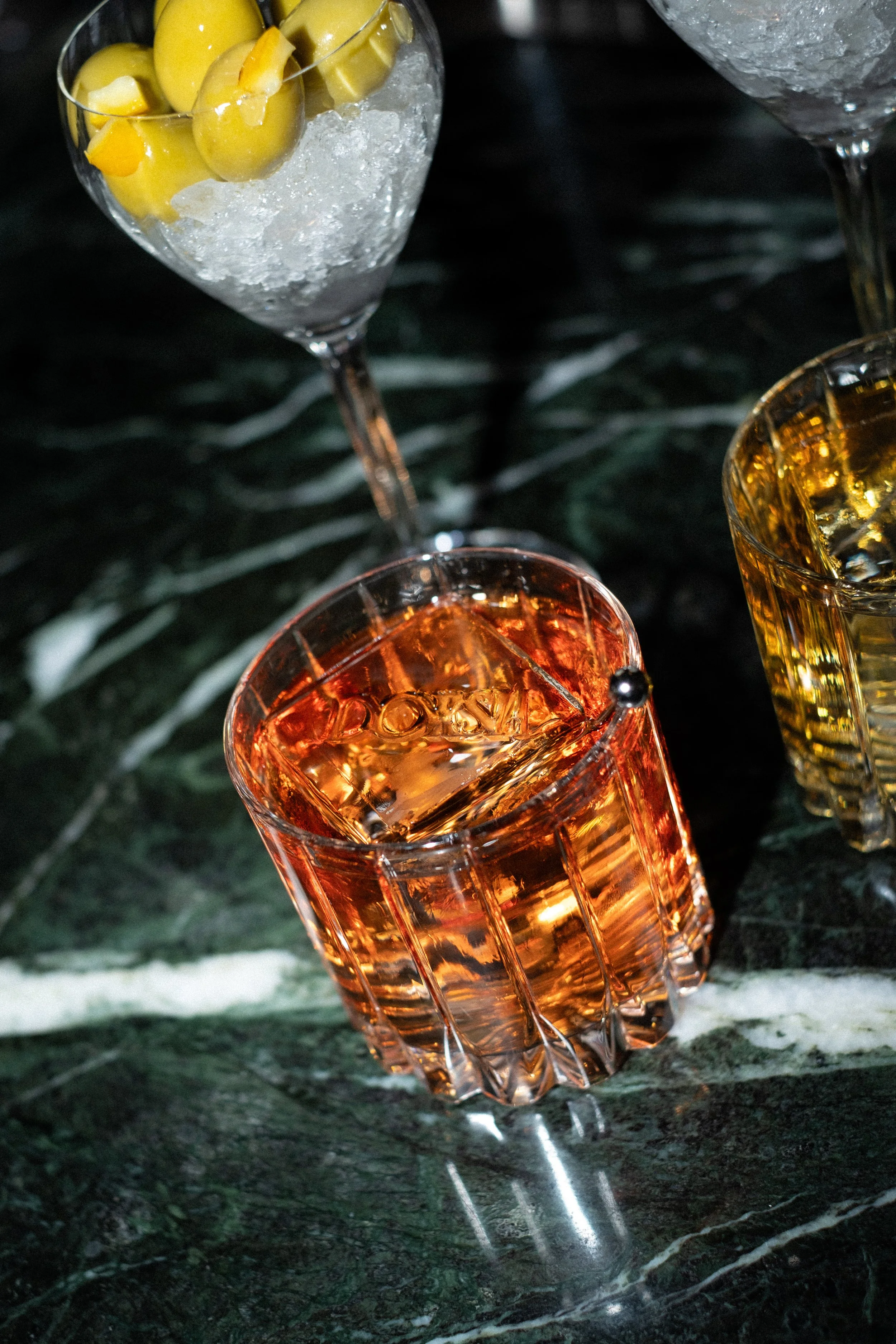 A glass of amber-colored liquor with ice cubes on a green marble surface, with other glasses in the background.