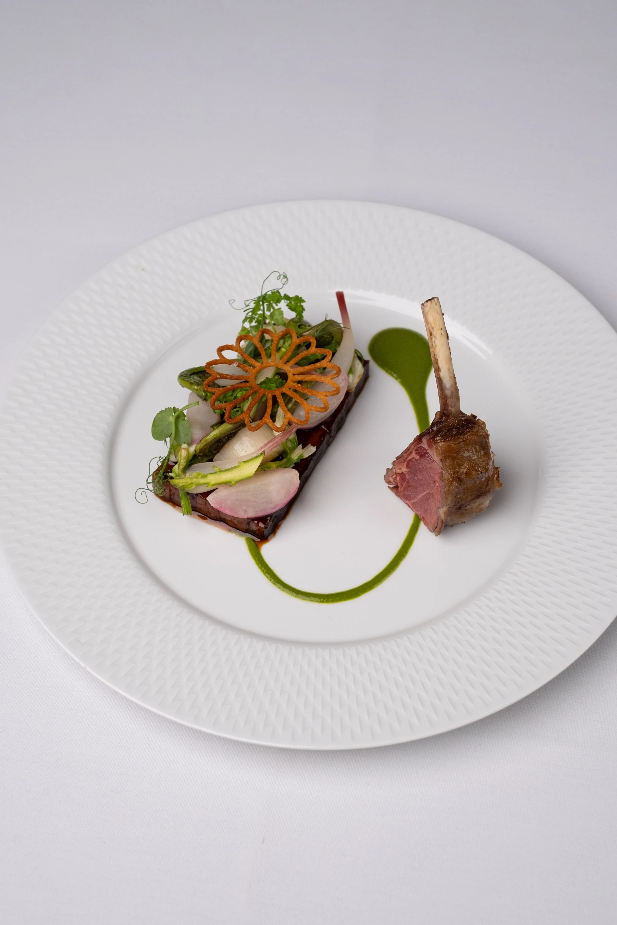 Gourmet dish with a cooked lamb chop, garnished with vegetables and a decorative vegetable chip, served with green sauce on a white plate.