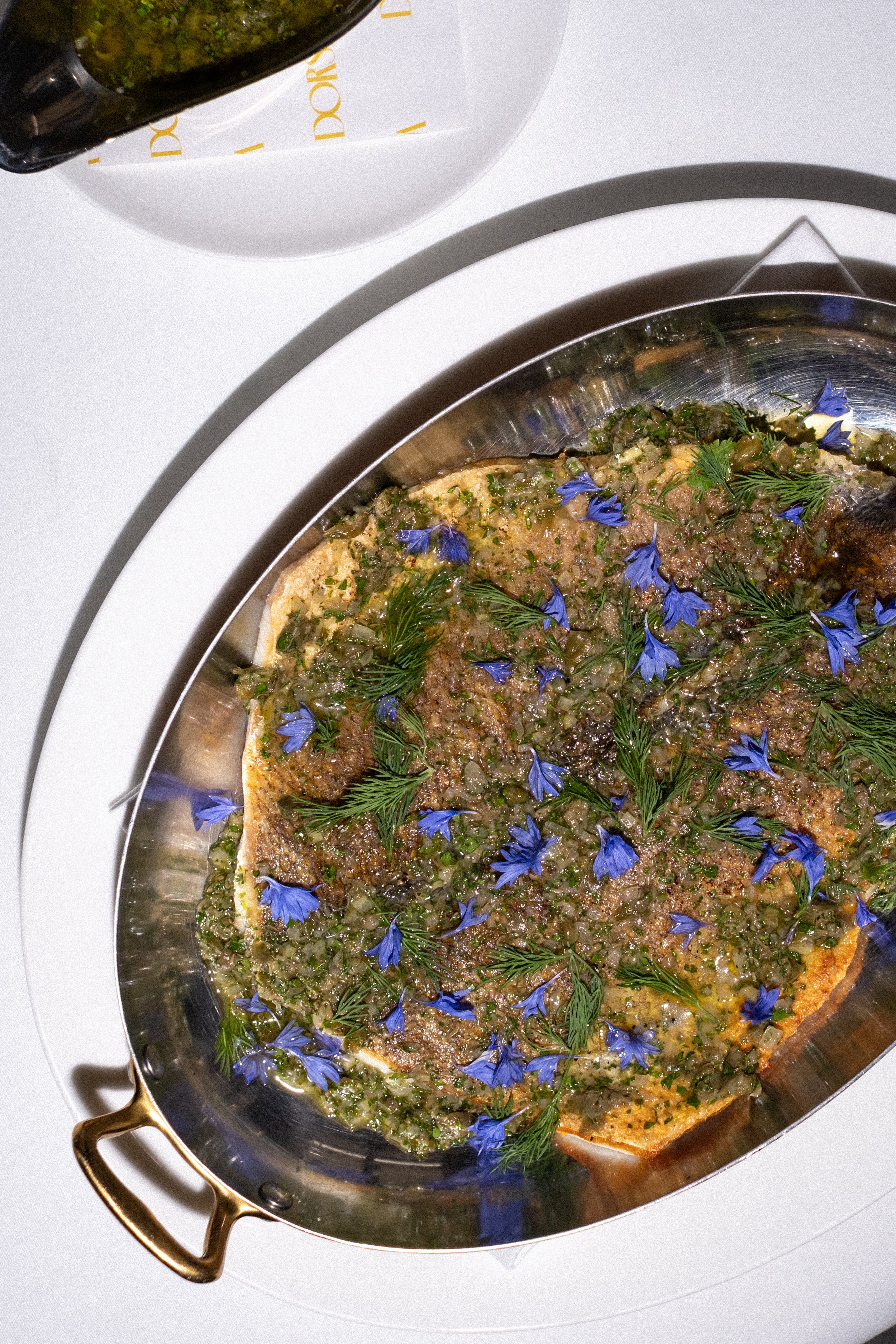 A fish fillet topped with chopped herbs, edible flowers, and garnished with dill, served on a round metal tray.