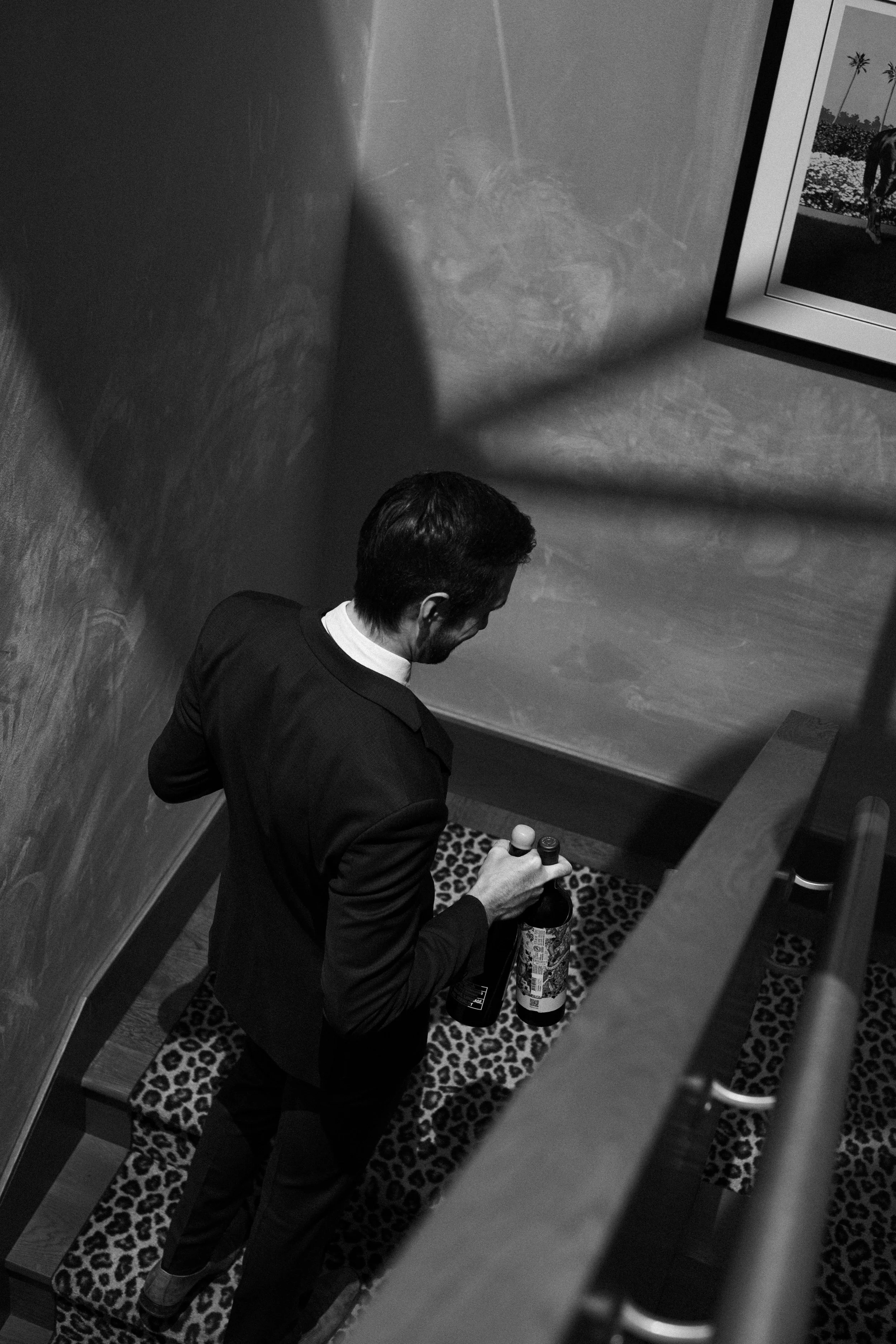 A man in a suit holding two bottles, standing on a leopard print rug at the top of a staircase.