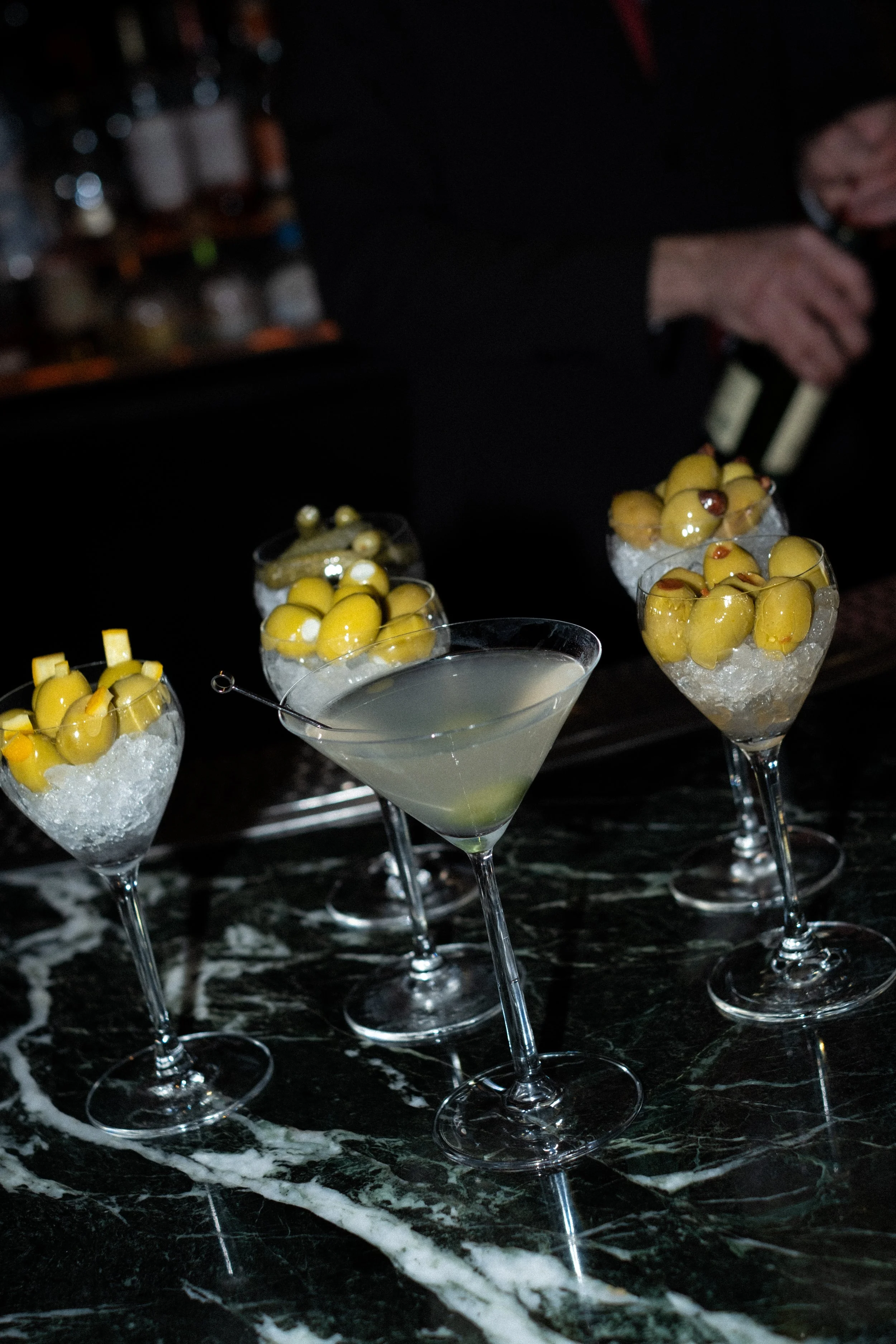 Four cocktail glasses with yellow olives on ice and a cocktail in the center, set on a dark marble bar counter.