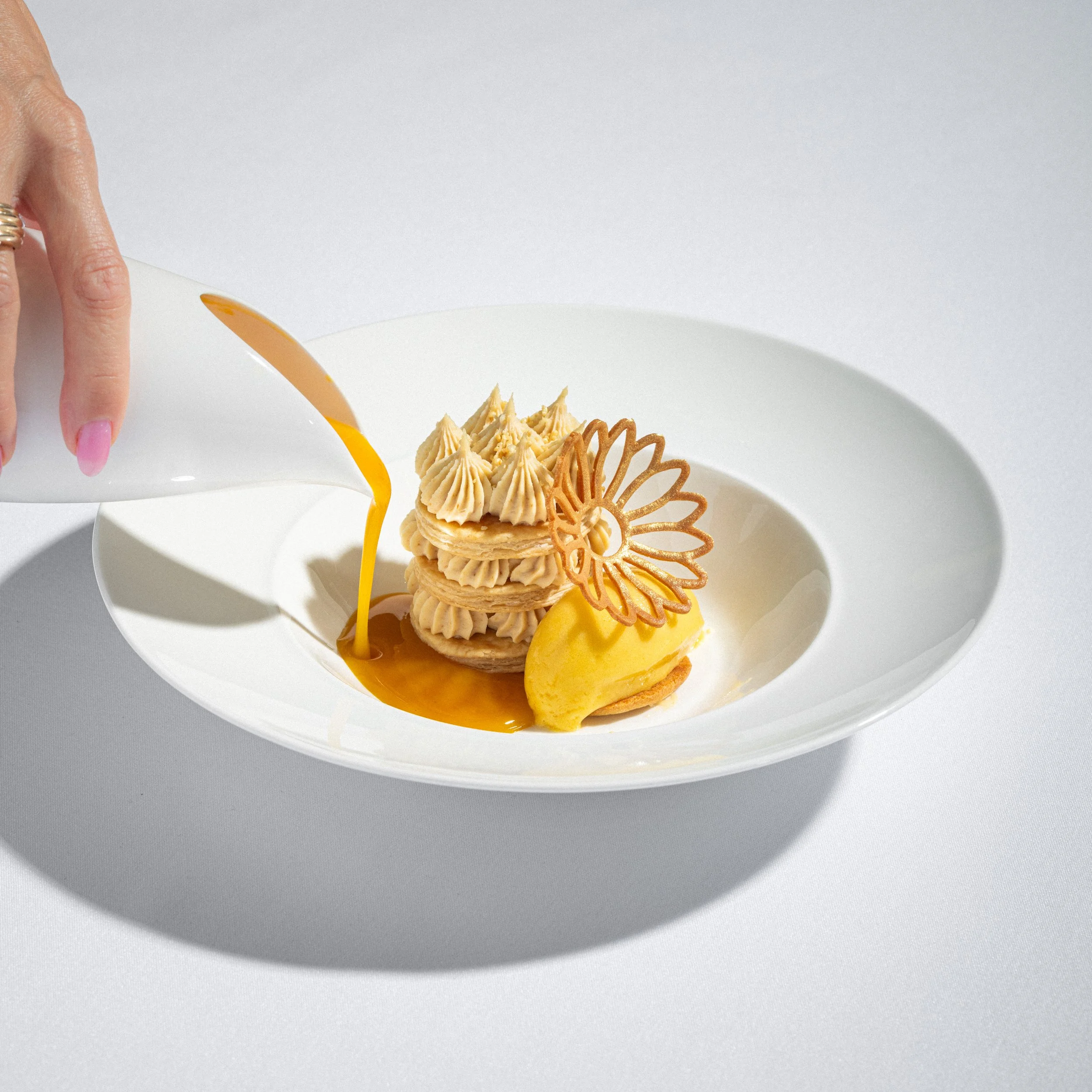 A white plate holding a layered dessert with piped cream, a decorative cookie, a lemon scoop, and a caramel sauce, with a hand pouring custard.