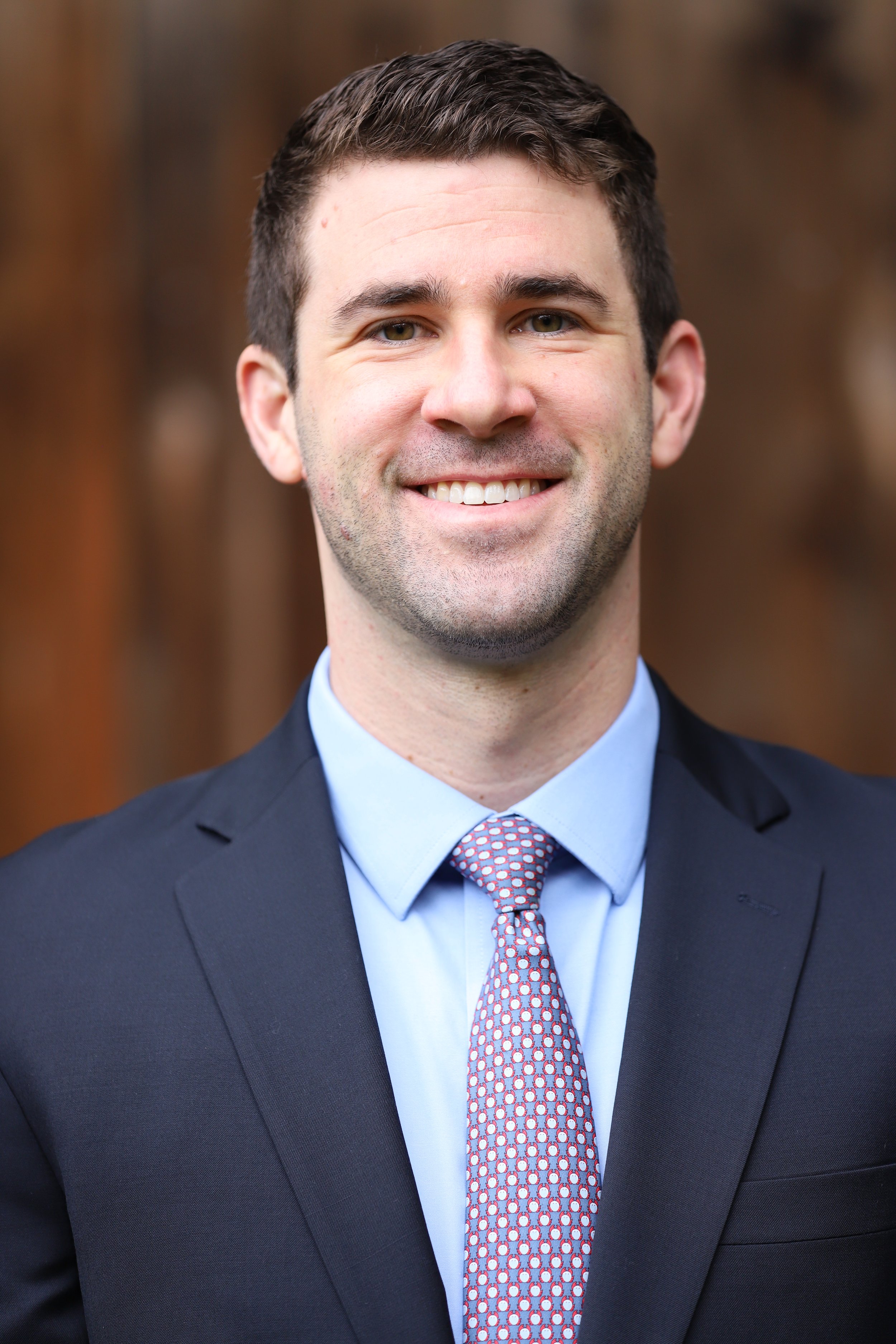 A portrait of a young man with short brown hair, dressed in a dark suit, light blue shirt, and a patterned tie, smiling at the camera. The background is blurred with warm tones.
