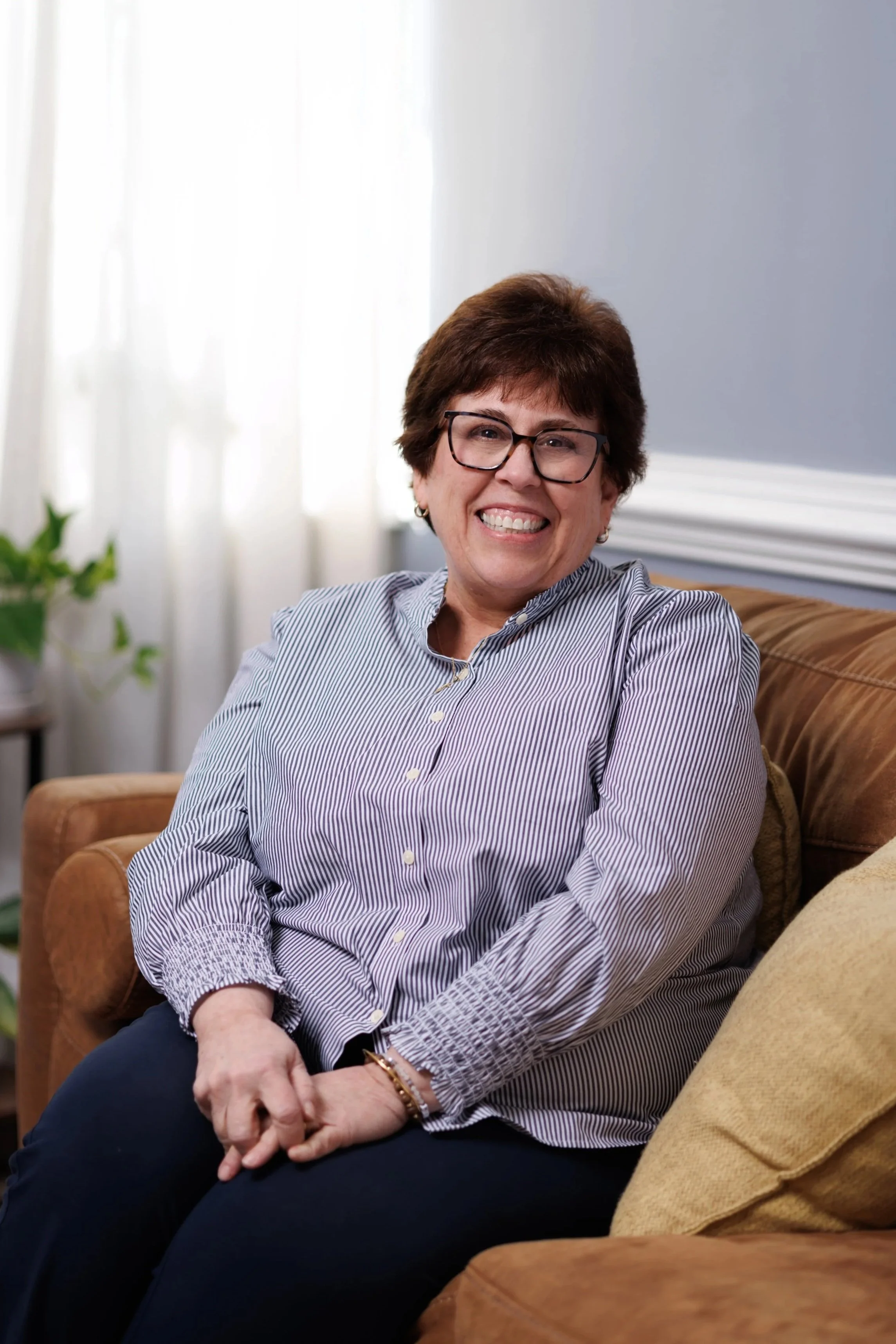 Maria Cunningham, Resident in Counseling, a smiling woman with short brown hair, wearing glasses, a striped button-up shirt, and dark pants, sitting on a brown couch in a well-lit room with a light-colored wall and curtains in the background.