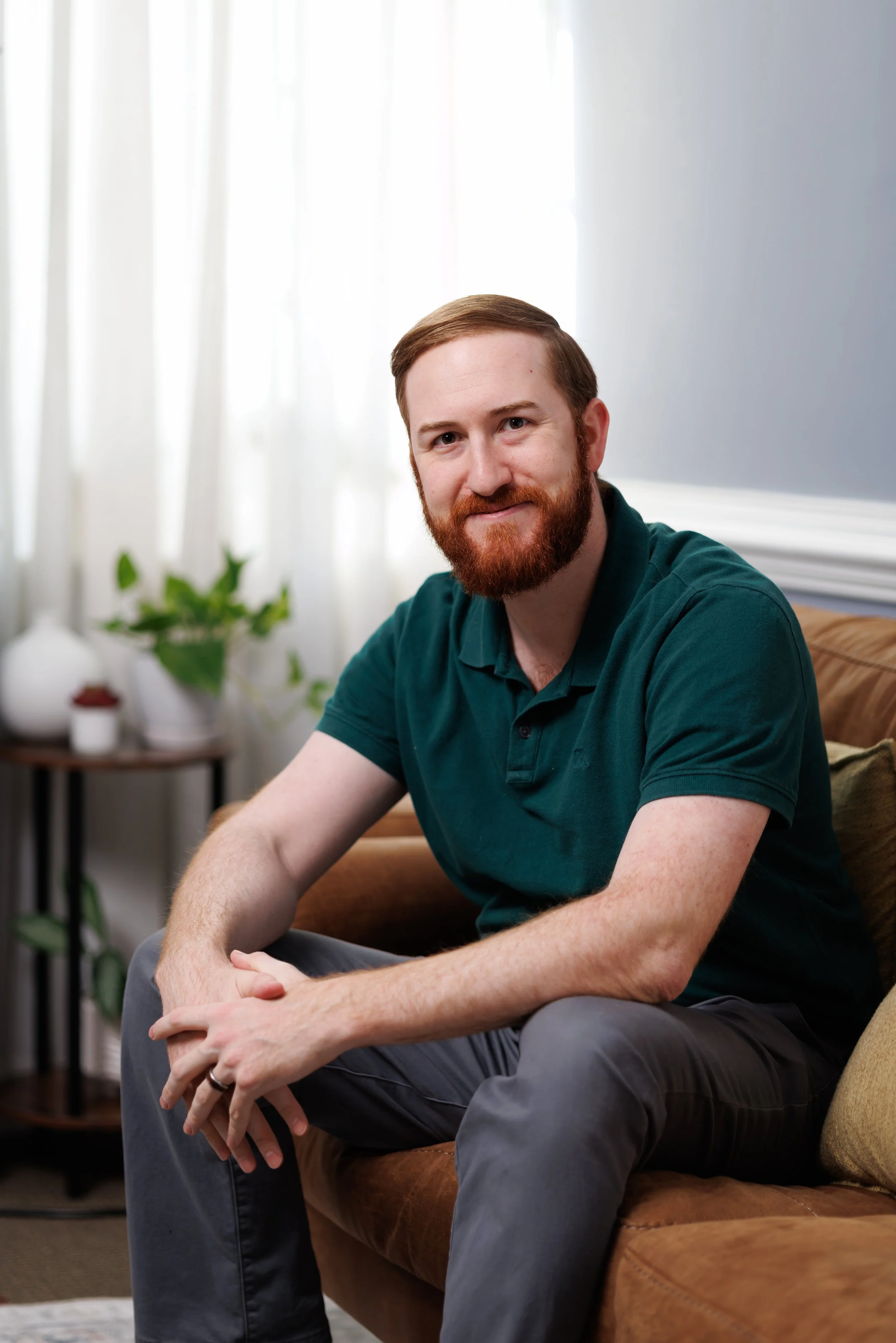 Dr. Christian Amalu, Psy.D., a man with a red beard and short hair sitting on a brown sofa in a living room, smiling at the camera. There are green plants and a side table with a white vase and a small pot in the background.
