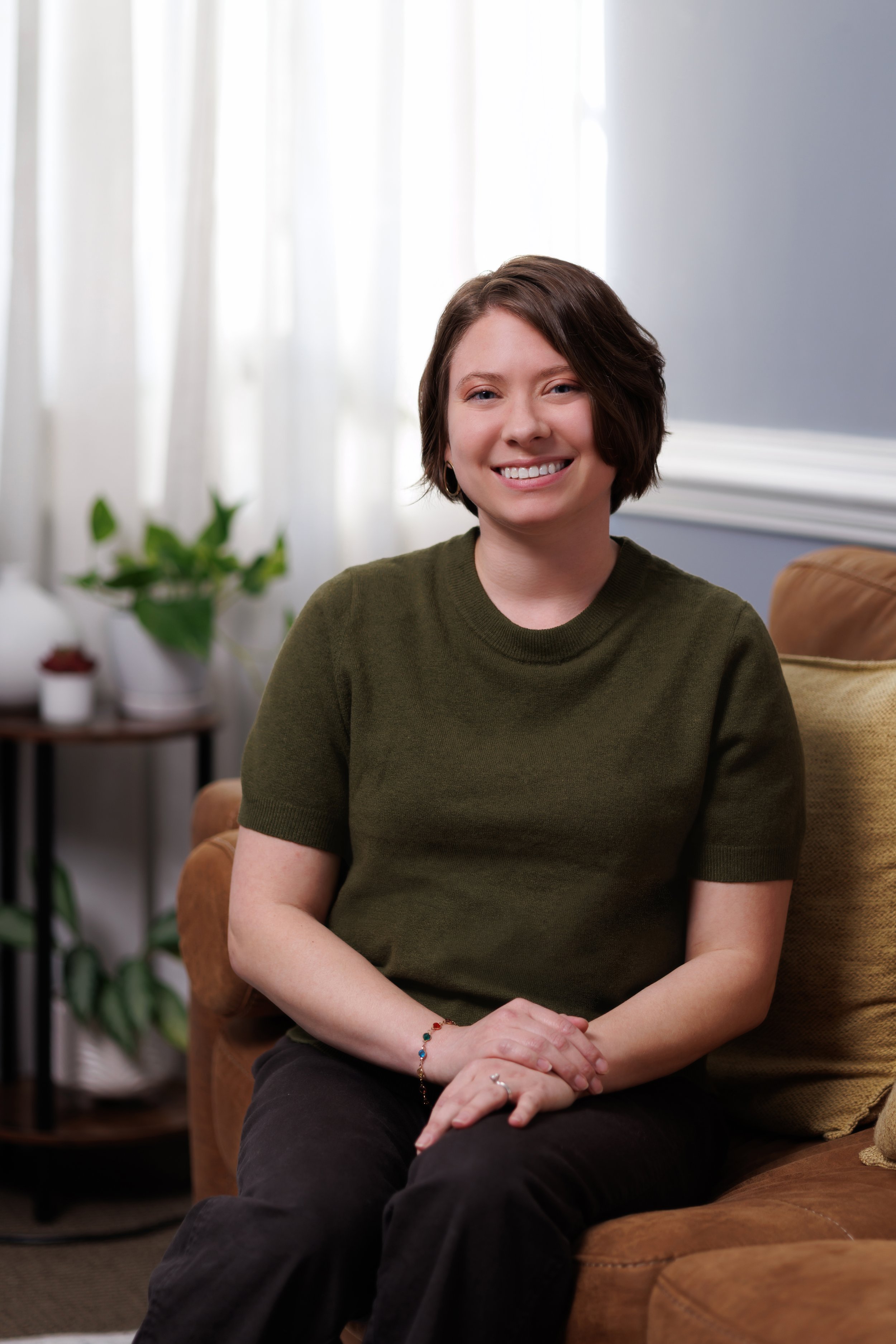 Katy Gustafson, LCPC, LPC, a woman with short brown hair smiling, sitting on a brown couch in a living room with a plant and side table in the background.