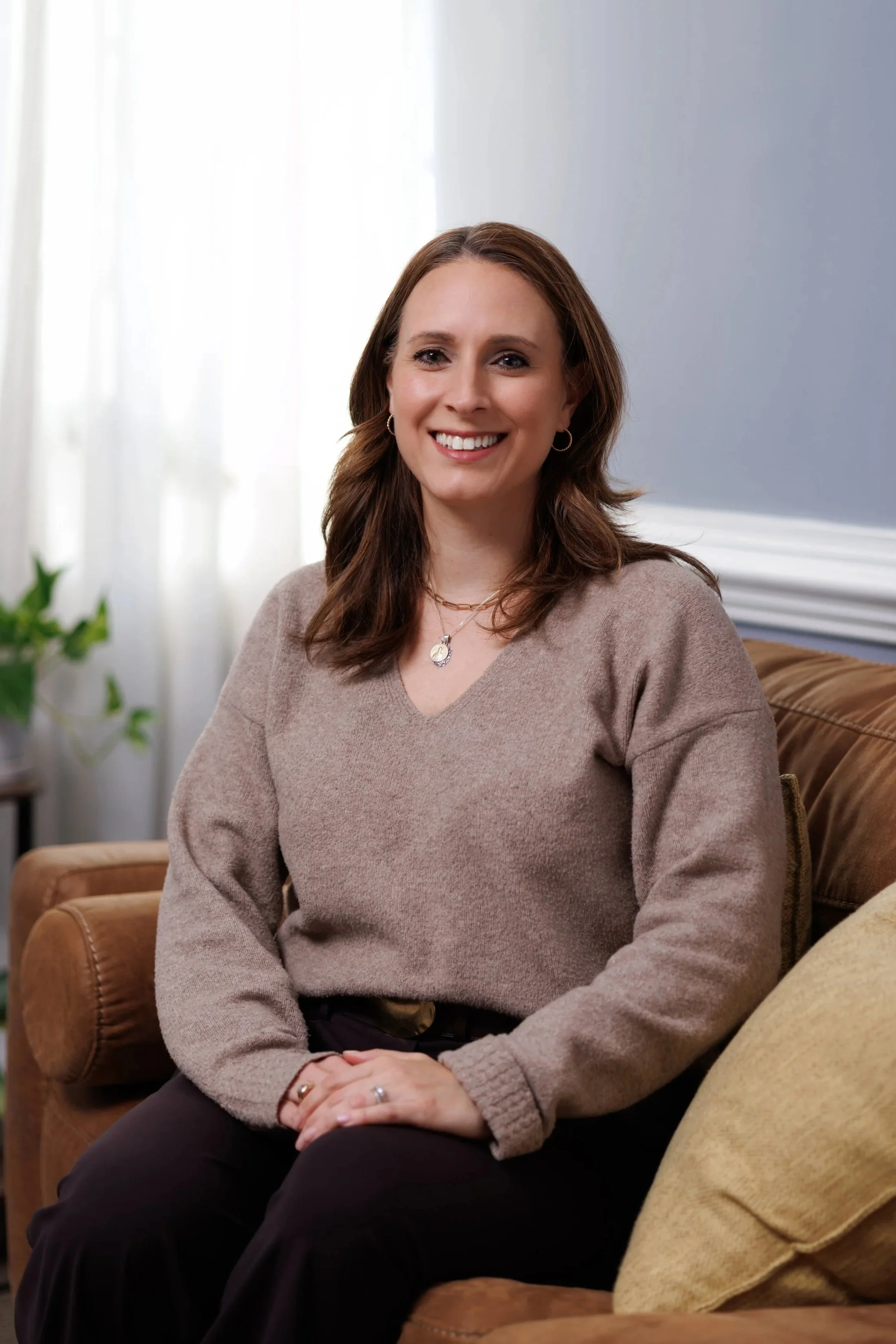 Katie Atwell, Resident in Counseling, a woman with brown hair, wearing a beige sweater and black pants, smiling while sitting on a brown couch in a bright room with a potted plant and white curtains in the background.