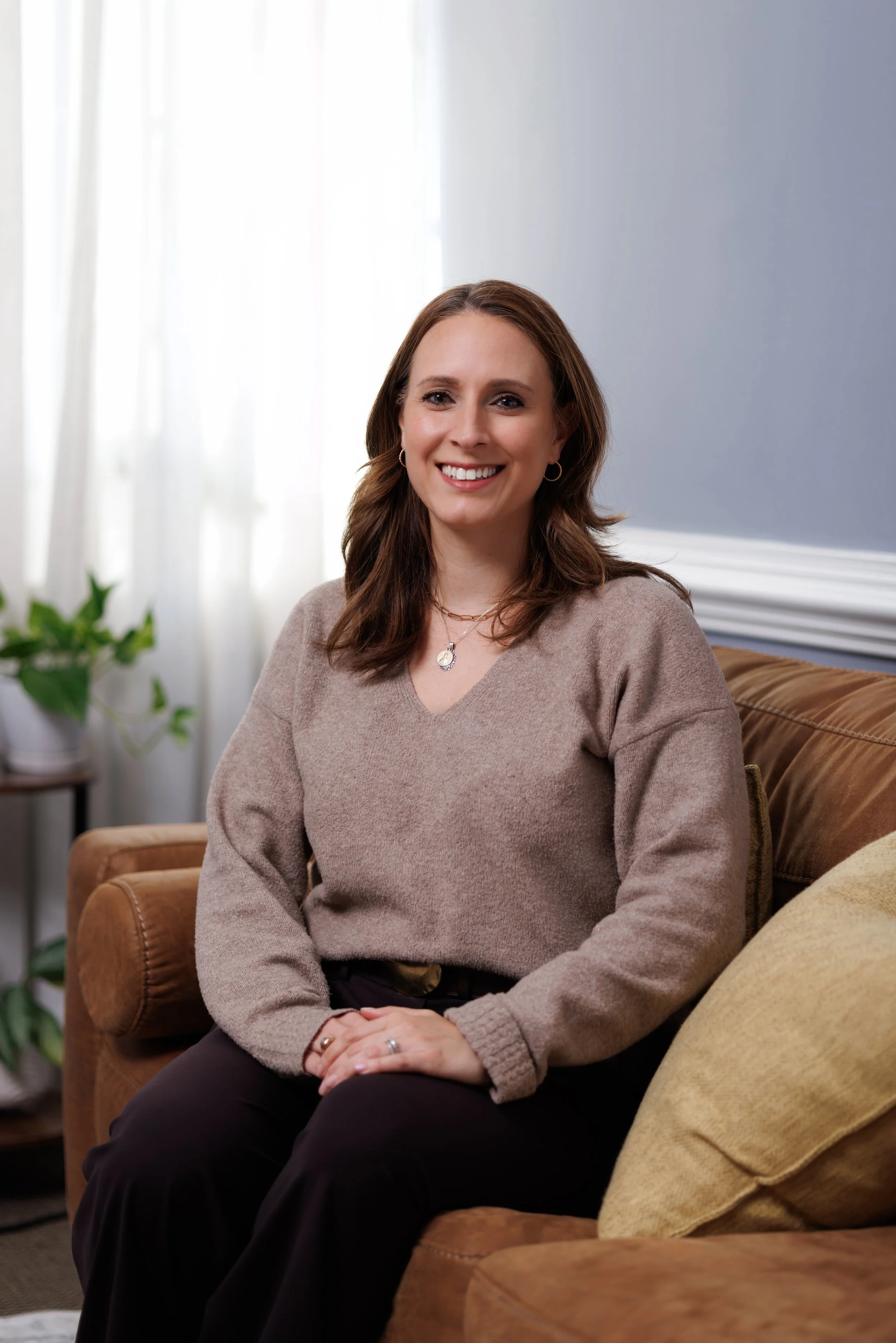 Katie Atwell, Resident in Counseling, a woman with brown hair, wearing a beige sweater and black pants, smiling while sitting on a brown couch in a bright room with a potted plant and white curtains in the background.