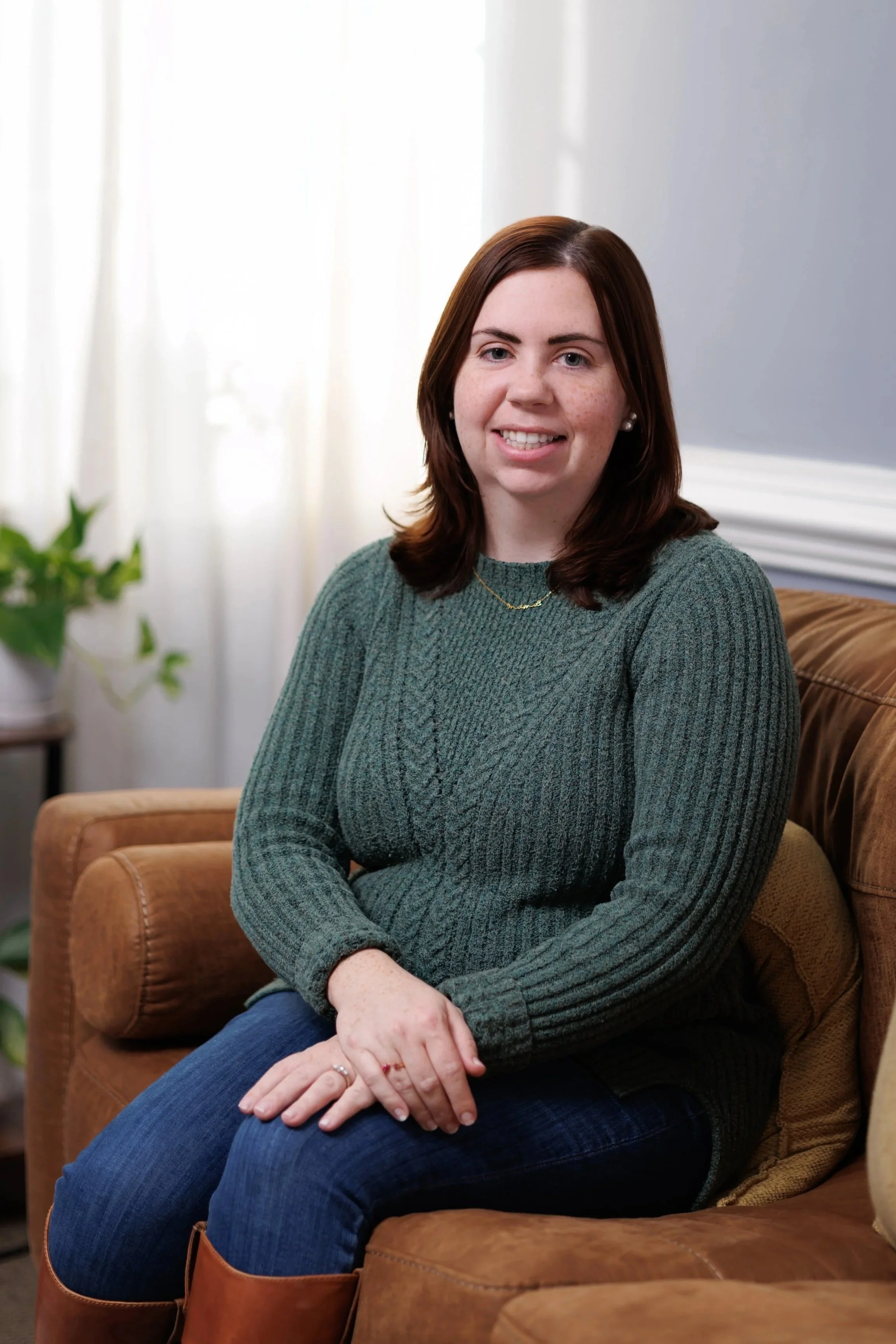 Catherine Boyle, Resident in Counseling, a woman with shoulder-length brown hair and fair skin sitting on a brown sofa in a well-lit room with white curtains, a small wooden table with a green plant, wearing a green knit sweater and blue jeans.
