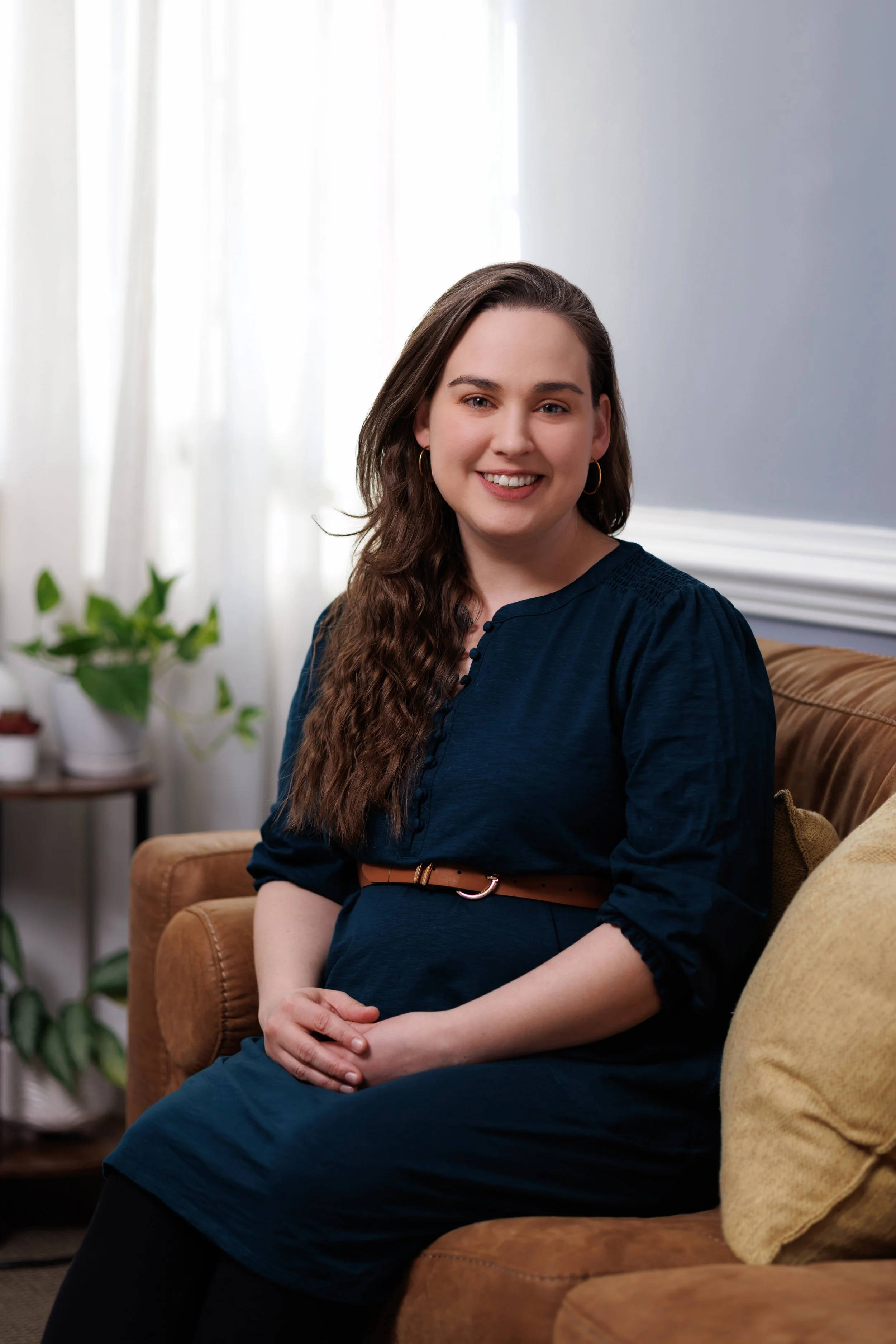 Micole Amalu, Counseling Intern, a woman with long, wavy brown hair, wearing a navy blue dress with a tan belt, sitting on a brown couch in a well-lit room with white curtains and houseplants in the background, smiling at the camera.