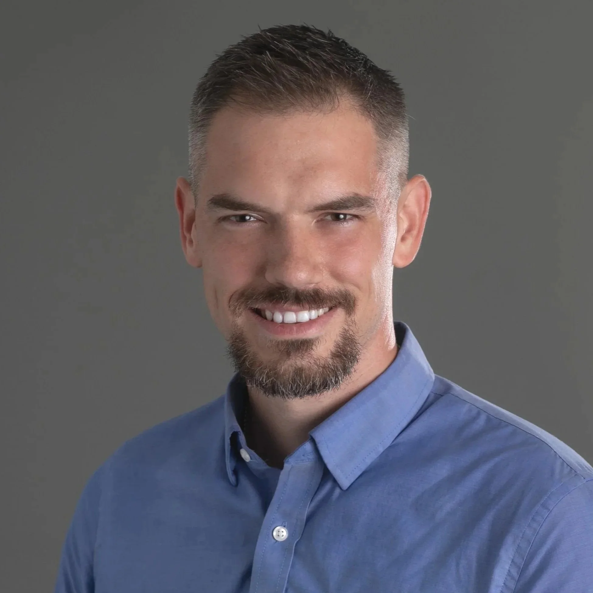 Jason Sparks, Resident in Counseling, who provides therapy for men. Headshot of a man with short brown hair, a beard, and a blue button-up shirt, smiling at the camera against a gray background.