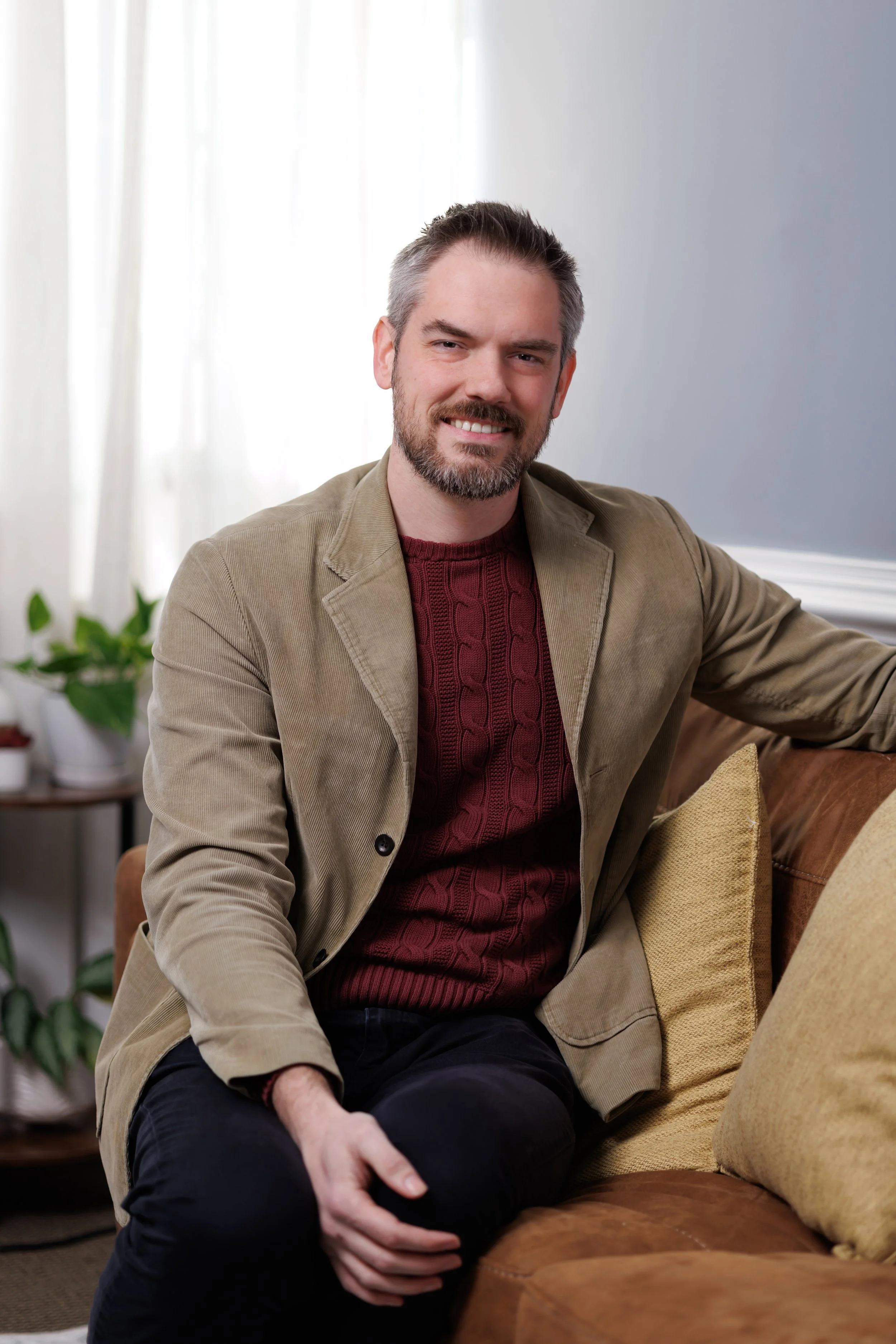 Jason Sparks, Resident in Counseling, a man with short dark hair and a beard, wearing a beige blazer and a maroon sweater, sitting on a brown leather couch with a beige pillow, smiling at the camera in a bright living room with a window and plants.