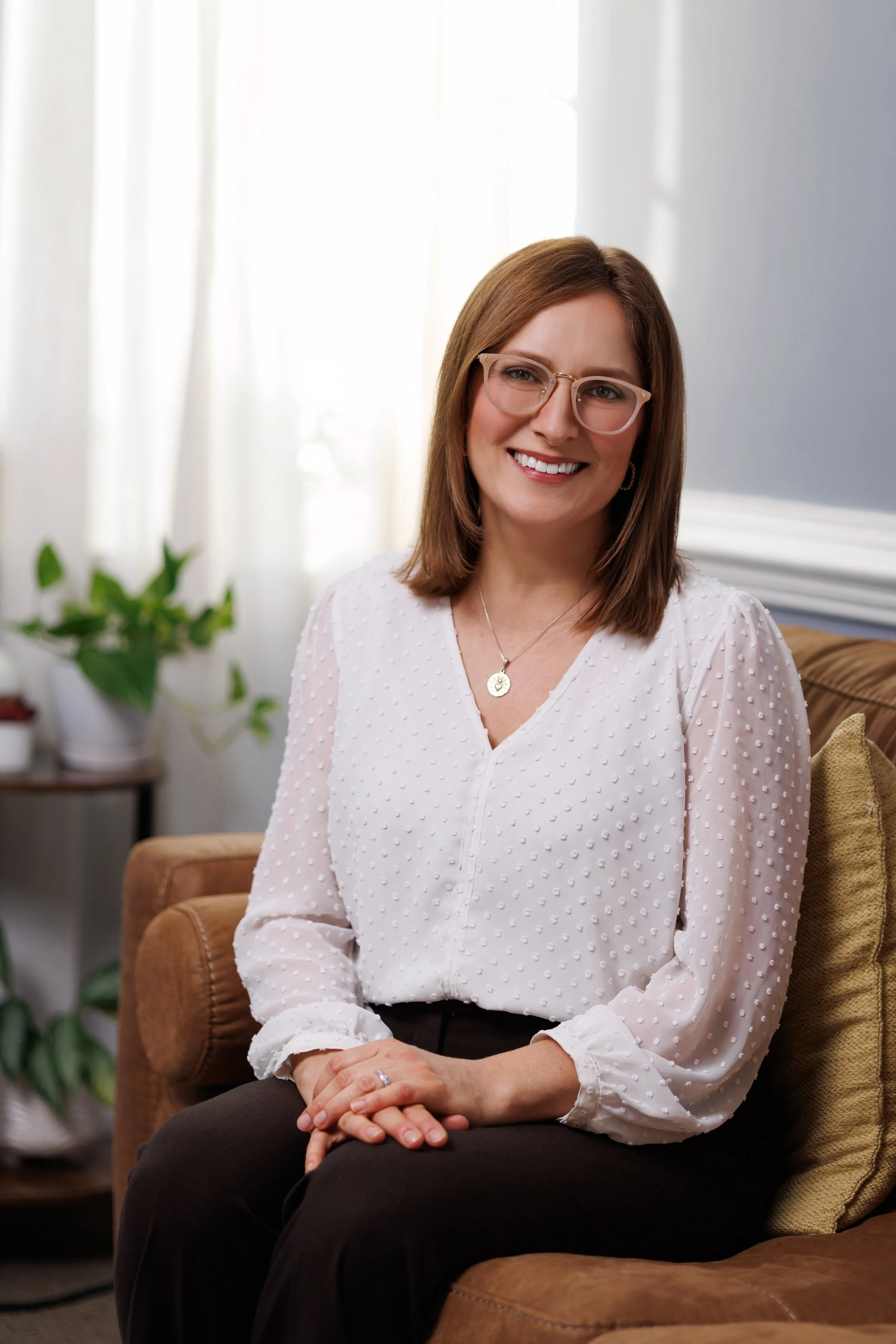 Irene Pruitt, LPC, a woman with brown hair and glasses, smiling, sitting on a brown couch inside a room with a window, plants, and a wall with blue and white trim.