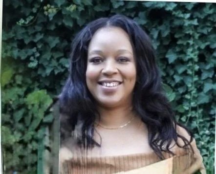 Woman with long, wavy black hair smiling outdoors in front of greenery and ivy.