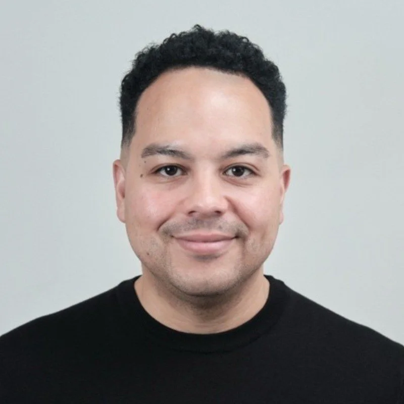 A smiling man with short curly black hair, wearing a black shirt, standing against a plain light gray background.