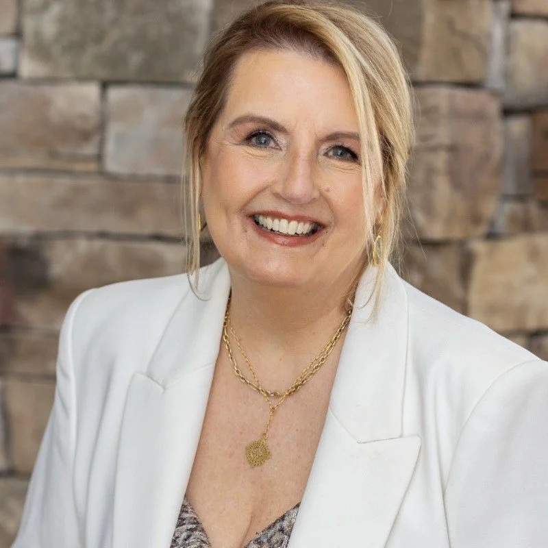 A smiling woman with blonde hair wearing a white blazer, gold jewelry, and a necklace with a pendant, standing against a stone wall background.