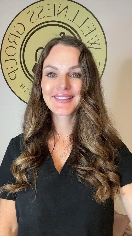 A woman with long, wavy brown hair and blue eyes smiling, wearing a black top, standing in front of a wall with a circular logo that reads "Wellness Group".