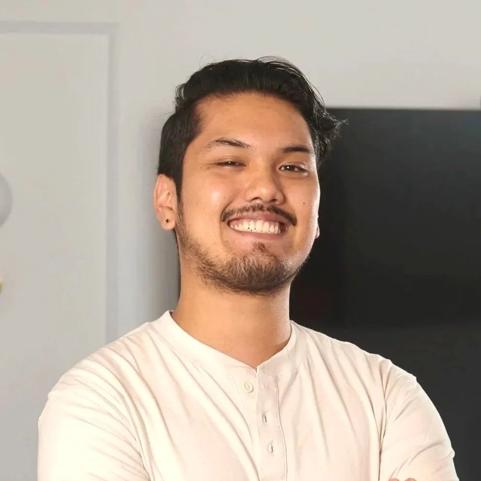 A young man with dark hair, facial hair, and earrings smiling with arms crossed, wearing a white shirt, indoors.