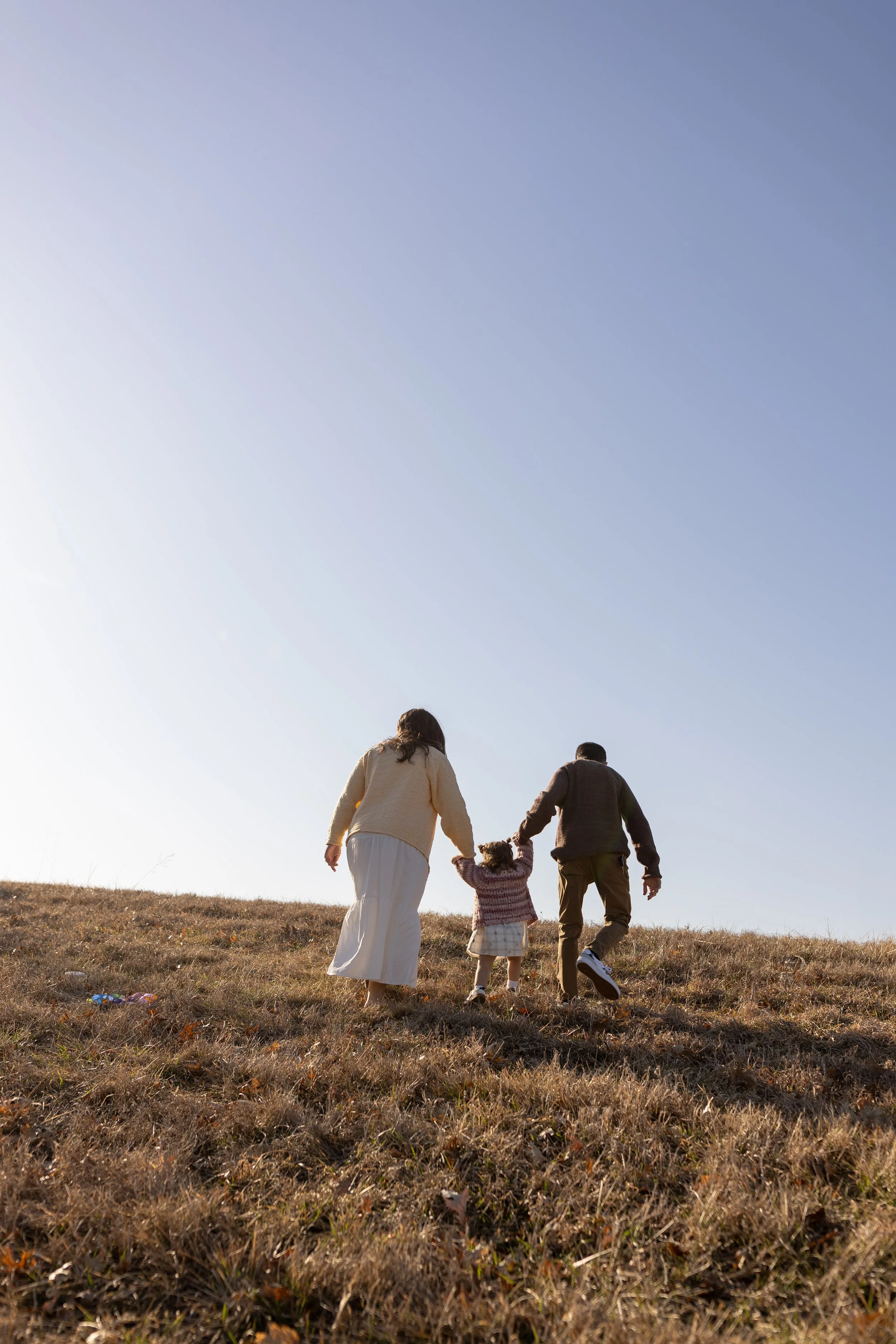 A family of three, including a woman, man, and young girl, walking up a grassy hill outdoors on a sunny day.