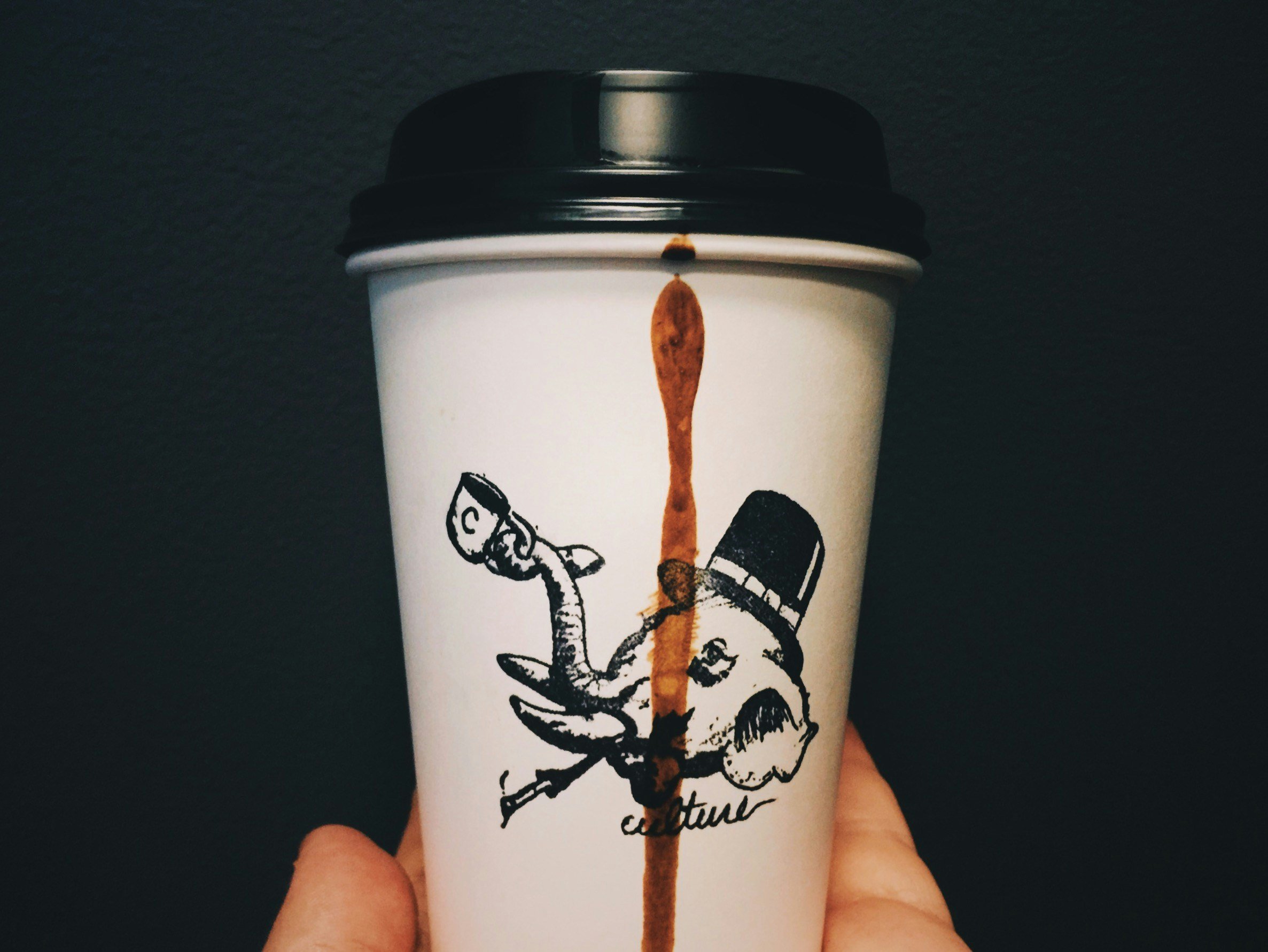 A white disposable coffee cup with black illustrations and handwriting, held by a person's hand. There is a streak of coffee dripping down the side of the cup.