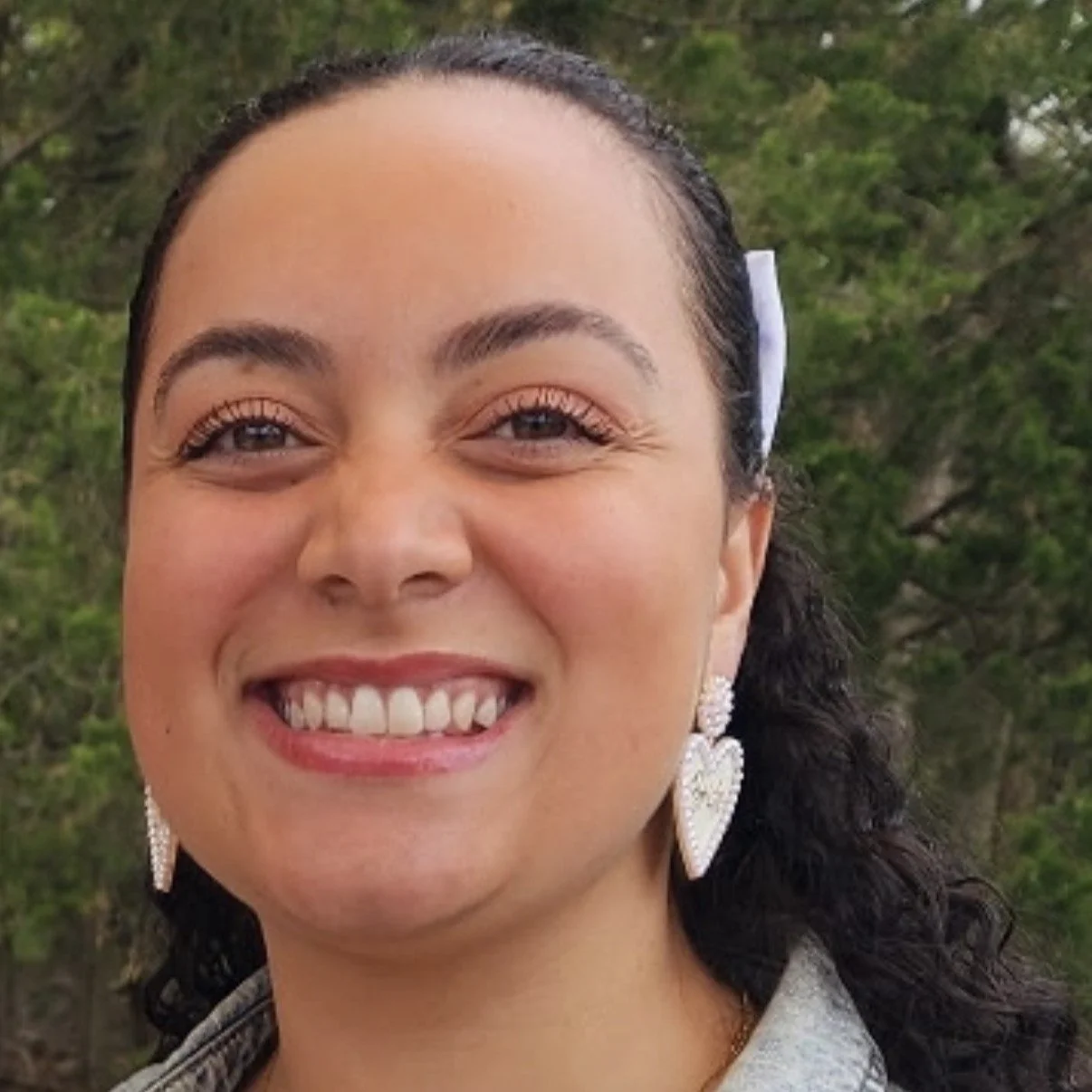 Close-up of a smiling woman with dark curly hair, wearing heart-shaped earrings, outdoors with green trees in the background.