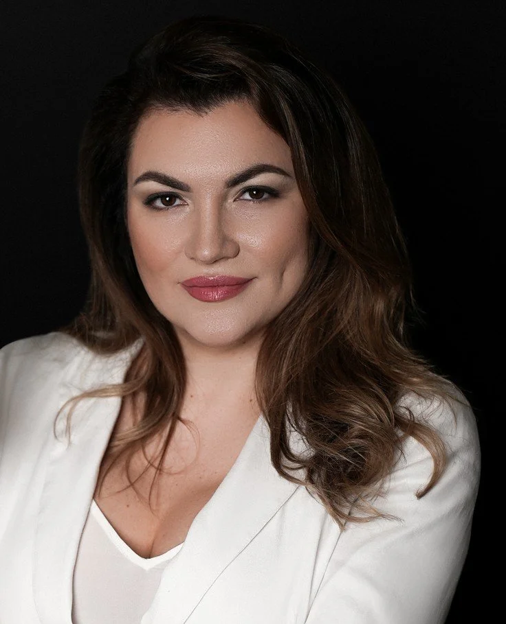 A woman with shoulder-length brown hair styled in loose waves, wearing a white blazer and smiling softly, against a black background.