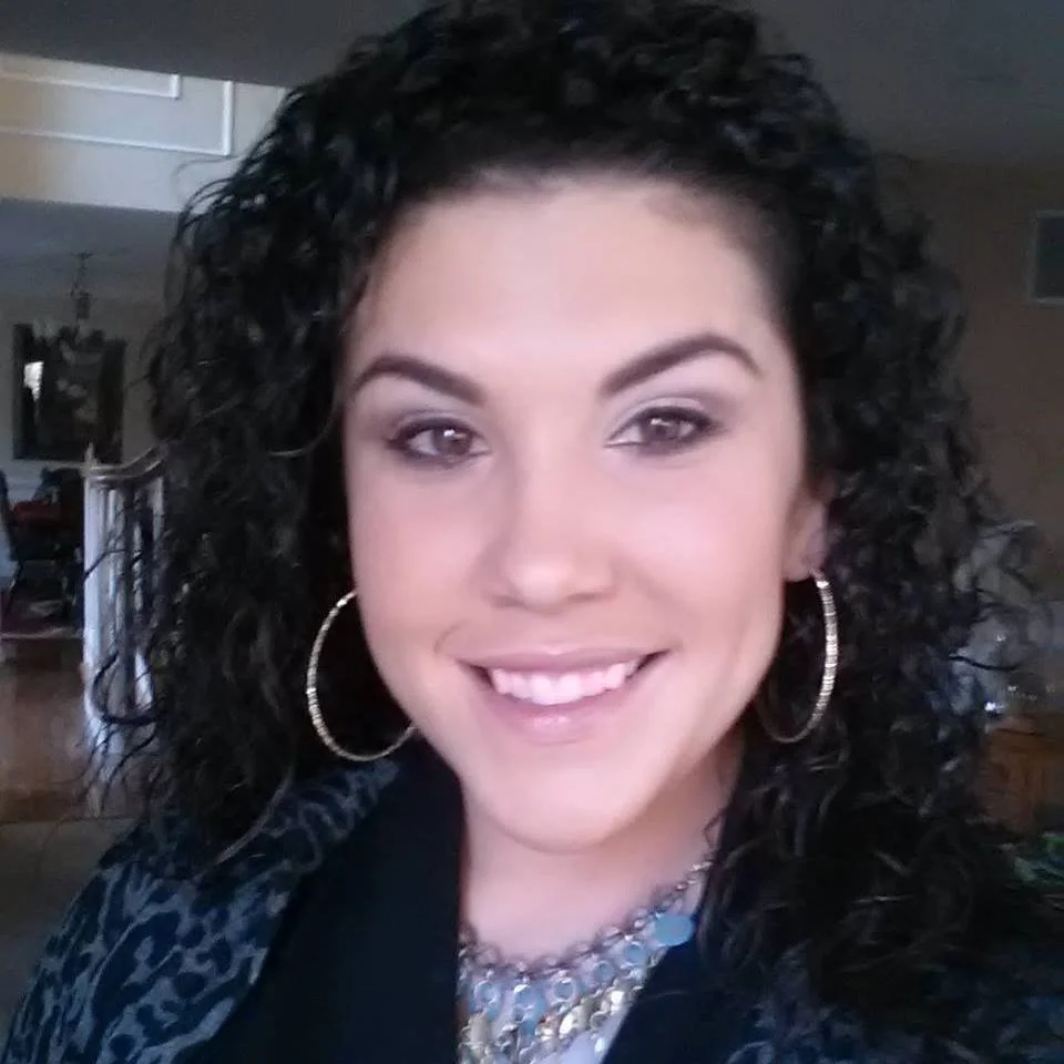 A woman with curly dark hair, smiling, wearing hoop earrings and a necklace, indoors with a blurred background.