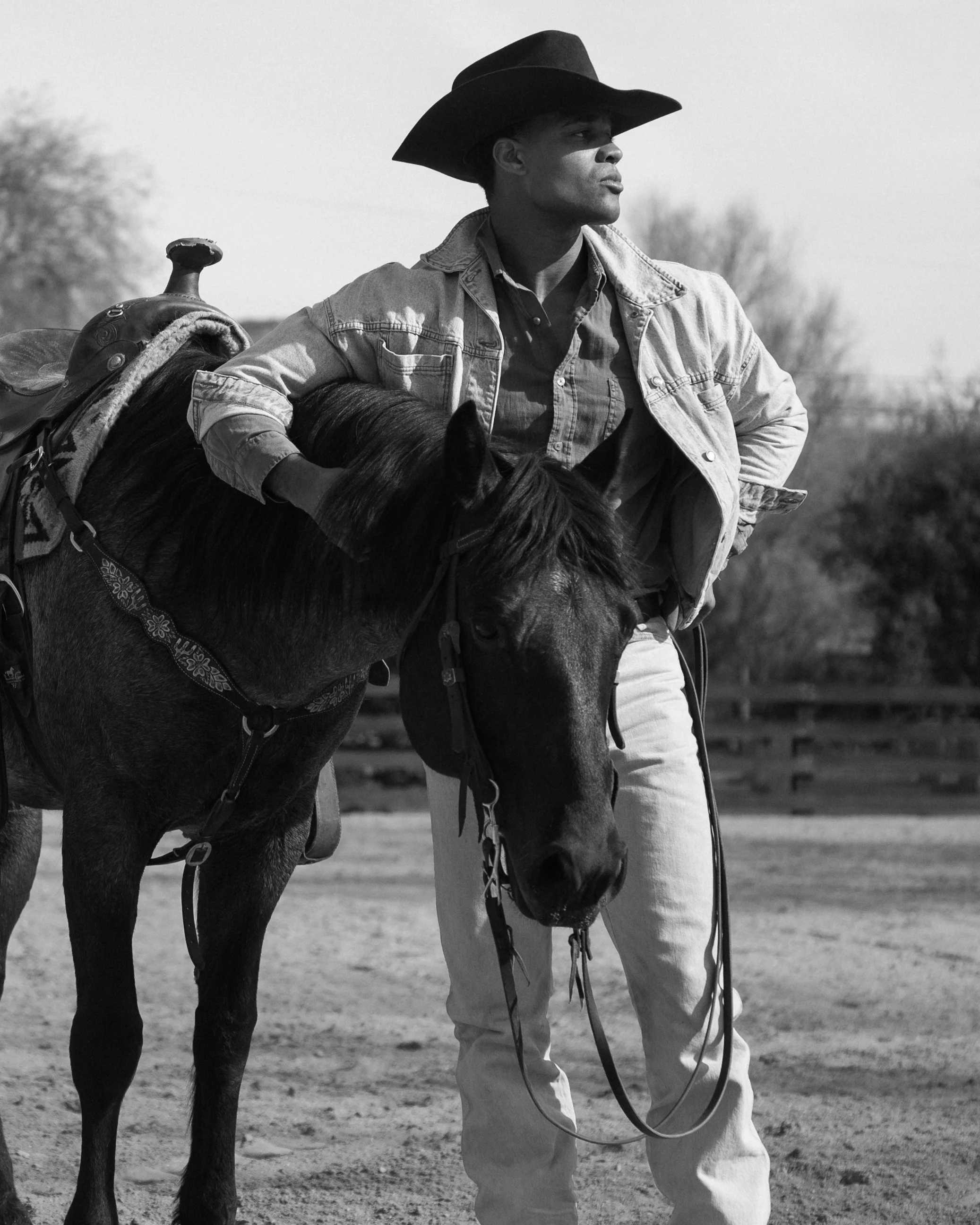 A man wearing a cowboy hat and denim jacket standing beside a horse on a dirt field, in a black and white photo.