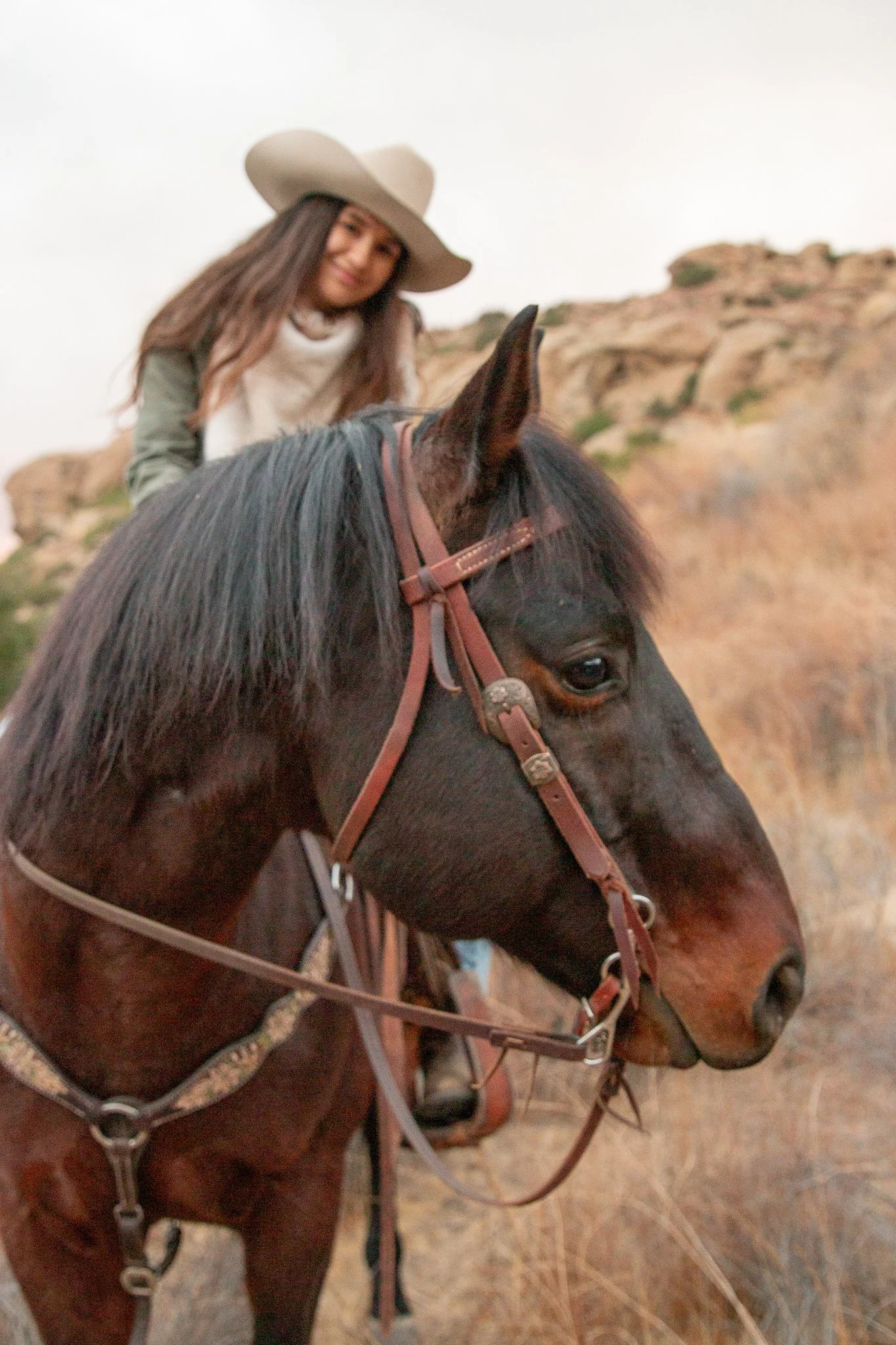 A woman with long hair, wearing a wide-brimmed hat, riding a dark brown horse with a brown leather bridle, in a desert-like landscape with rocky formations in the background.