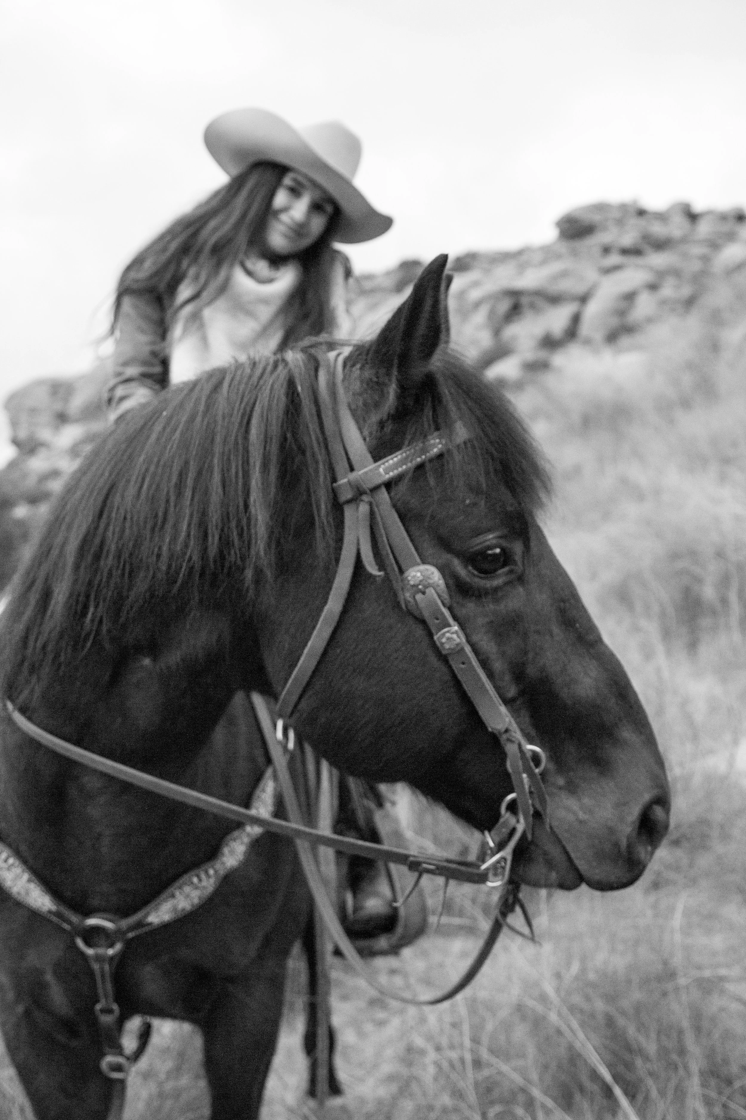 A woman riding a horse outdoors in a natural landscape, wearing a wide-brimmed hat and smiling.