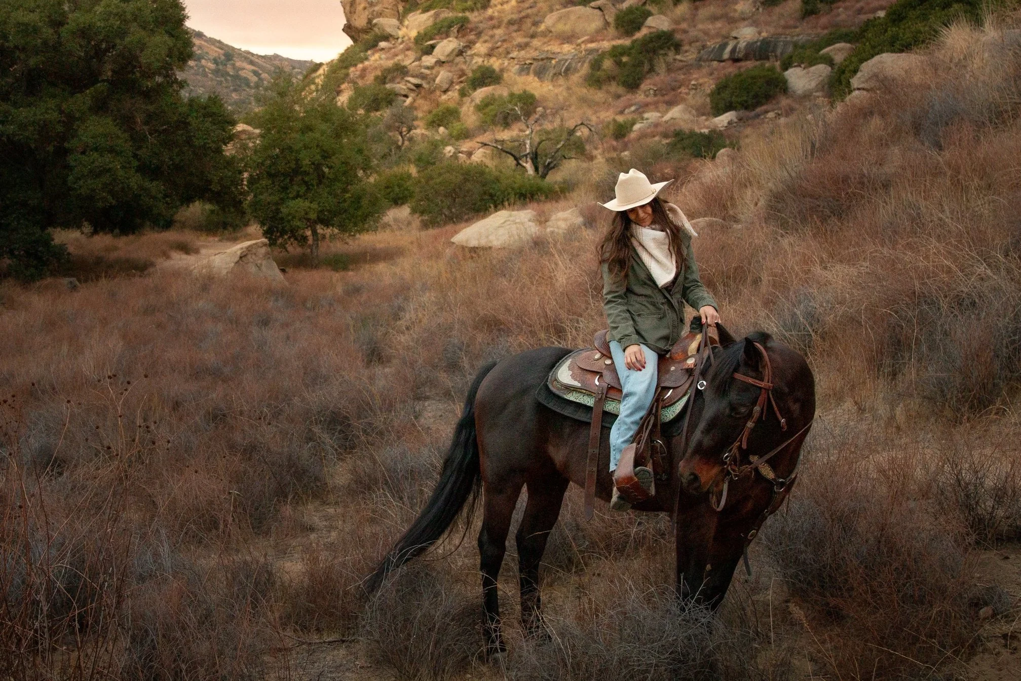 A woman riding a horse through a dry, mountainous landscape with sparse vegetation and rocks during sunset.