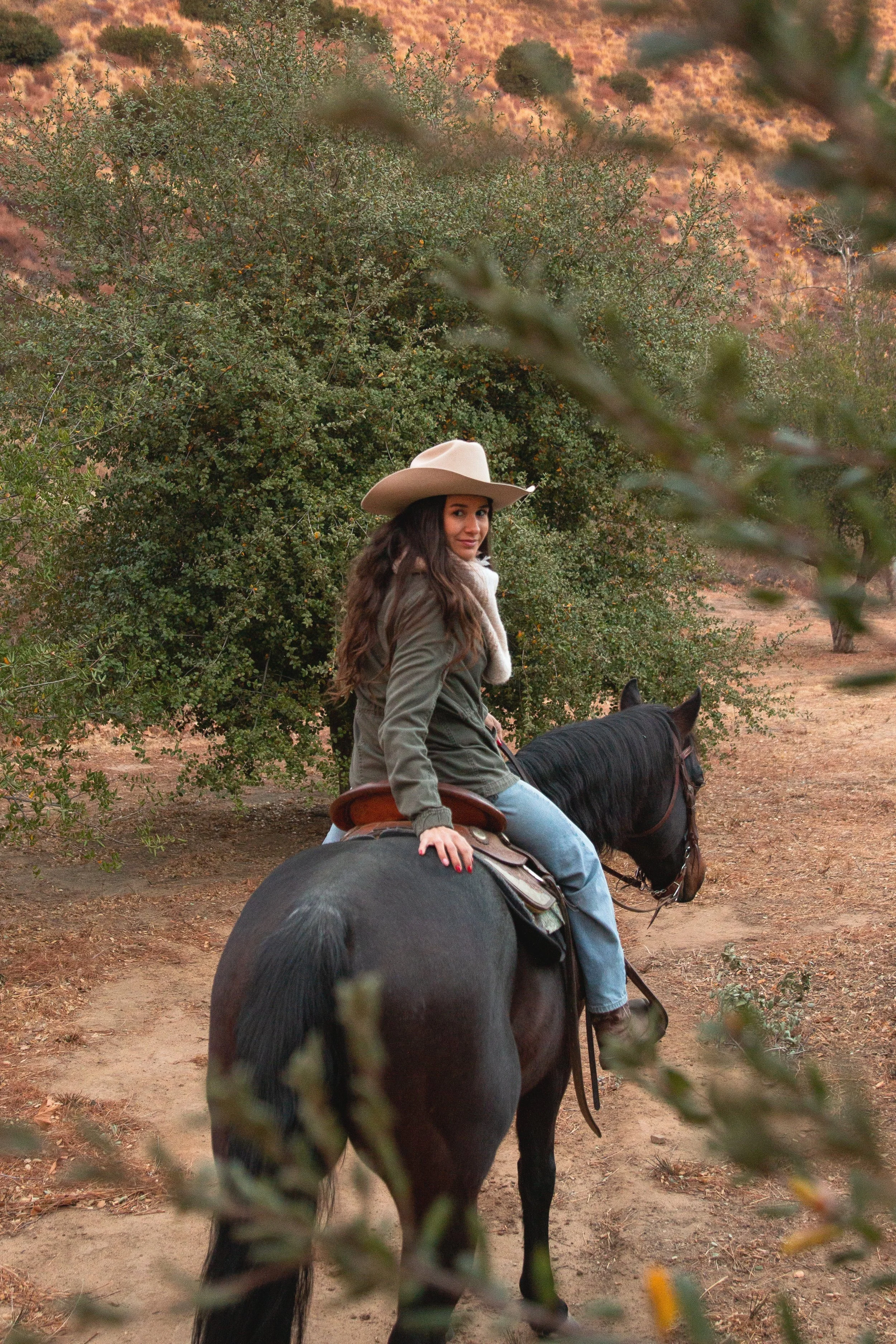 A woman with long hair, wearing a wide-brimmed hat and a green jacket, is riding a black horse in a natural outdoor setting with bushes and trees.