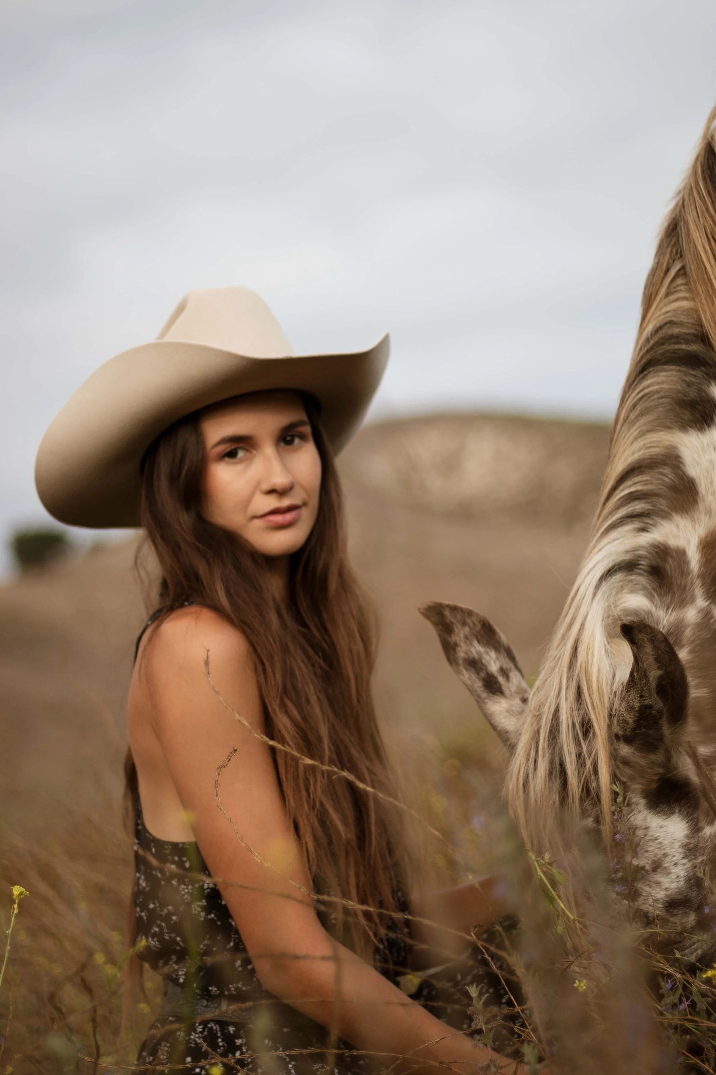 A woman with long brown hair wearing a beige cowboy hat and a dark dress with a floral pattern, sitting in a field with tall grass and a horse grazing nearby.