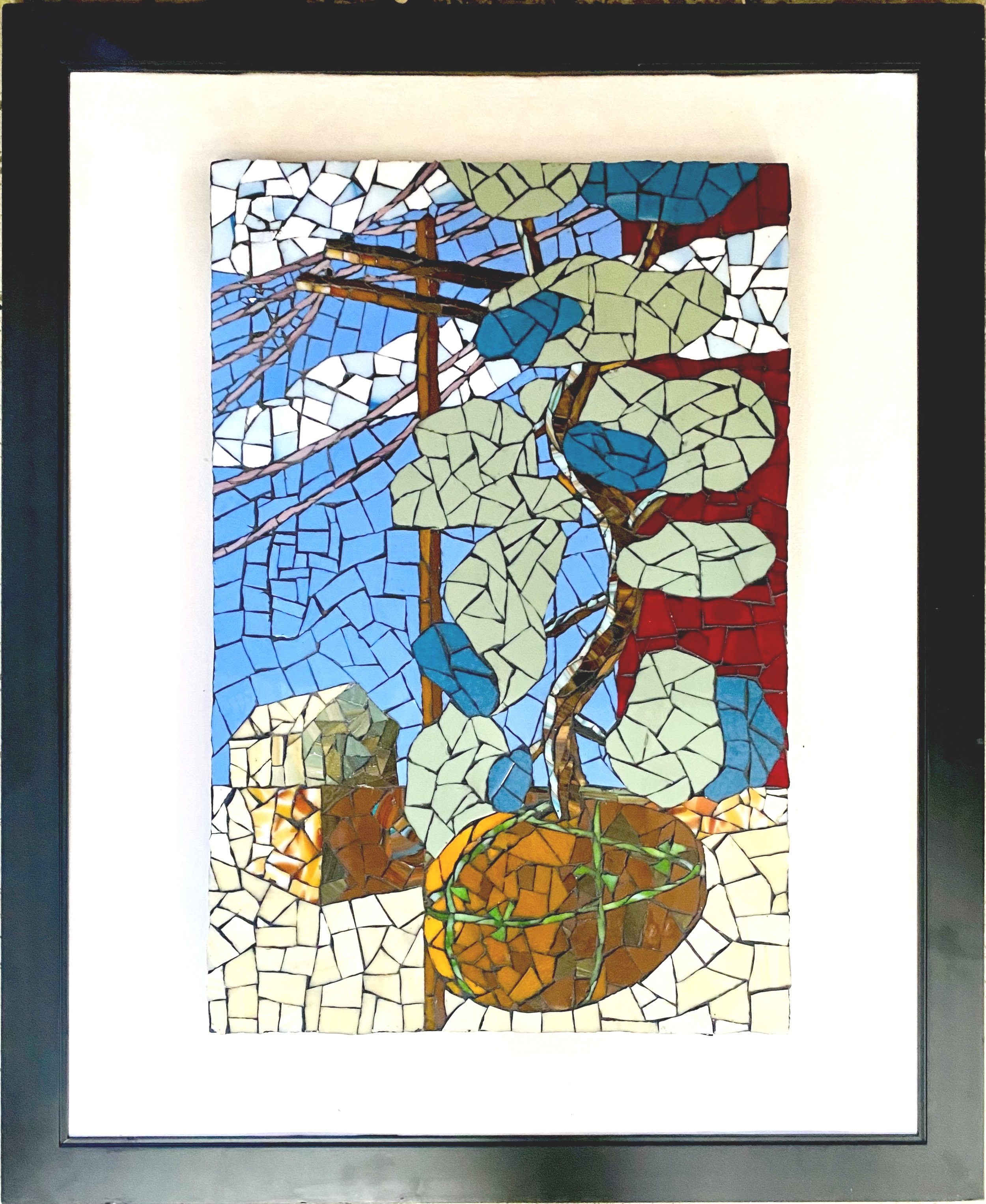 Framed glass mosaic of a potted tree with large green and blue leaves, set against a sky with power lines, clouds, and surrounding buildings