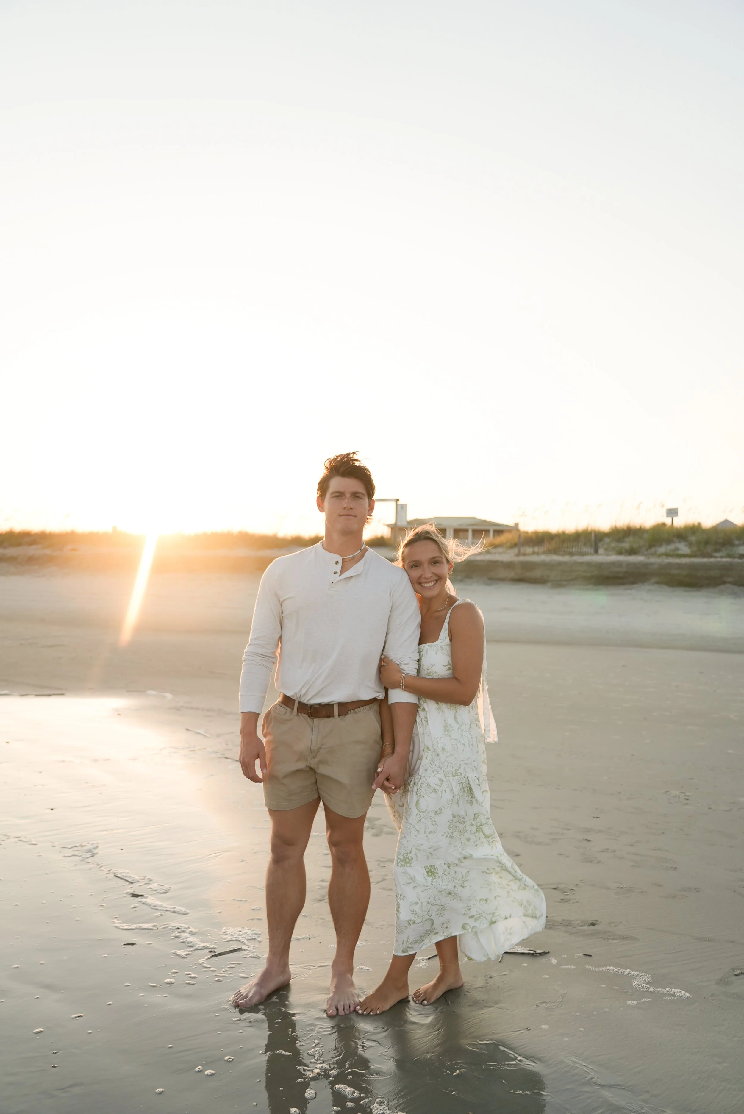 A young couple standing on the beach at sunset, holding hands and smiling, with the ocean in the background.