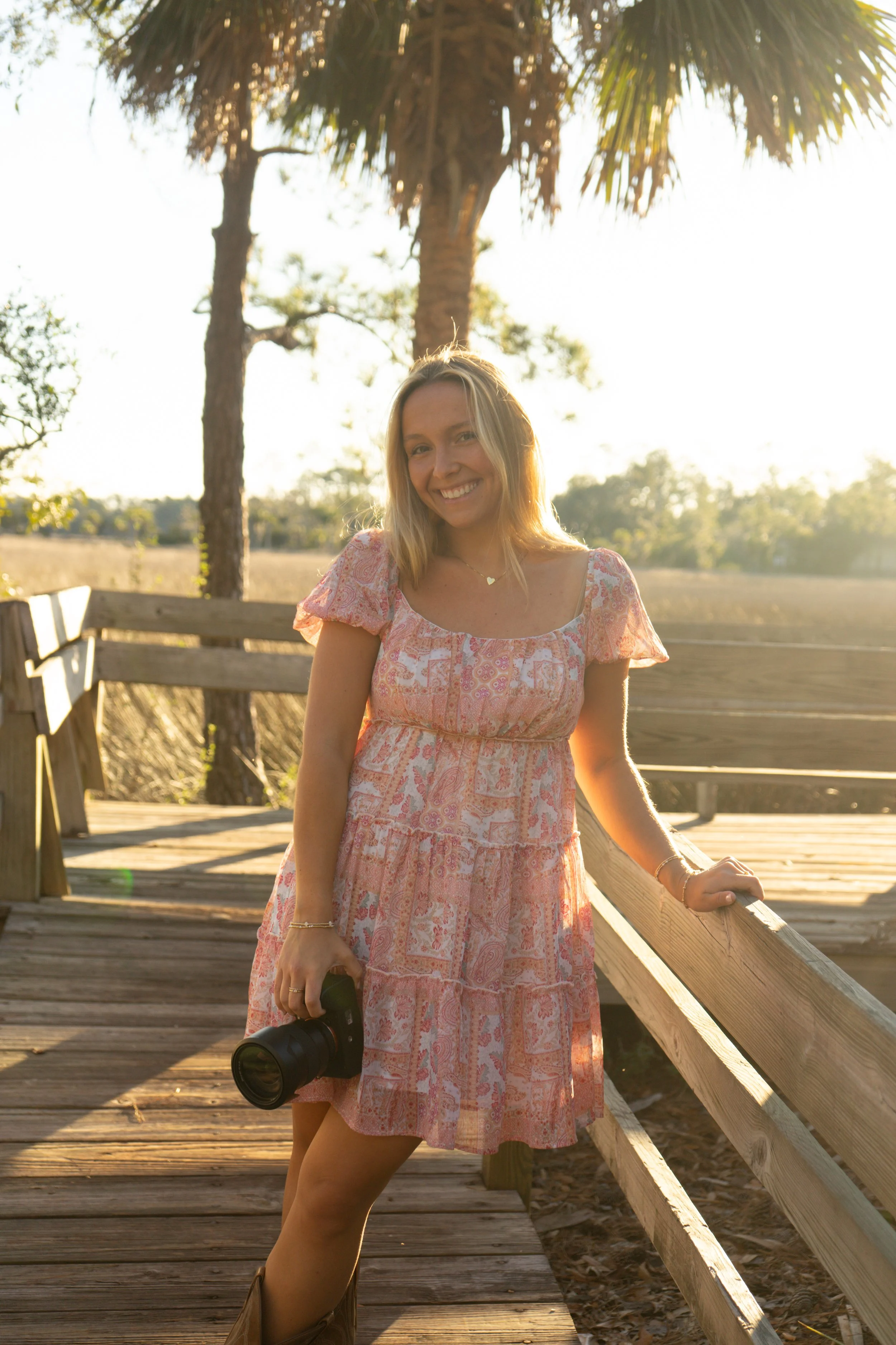 A young woman with blonde hair smiling, wearing a pink patterned dress, standing on a wooden pathway holding a camera, with a tree and field in the background during sunset.