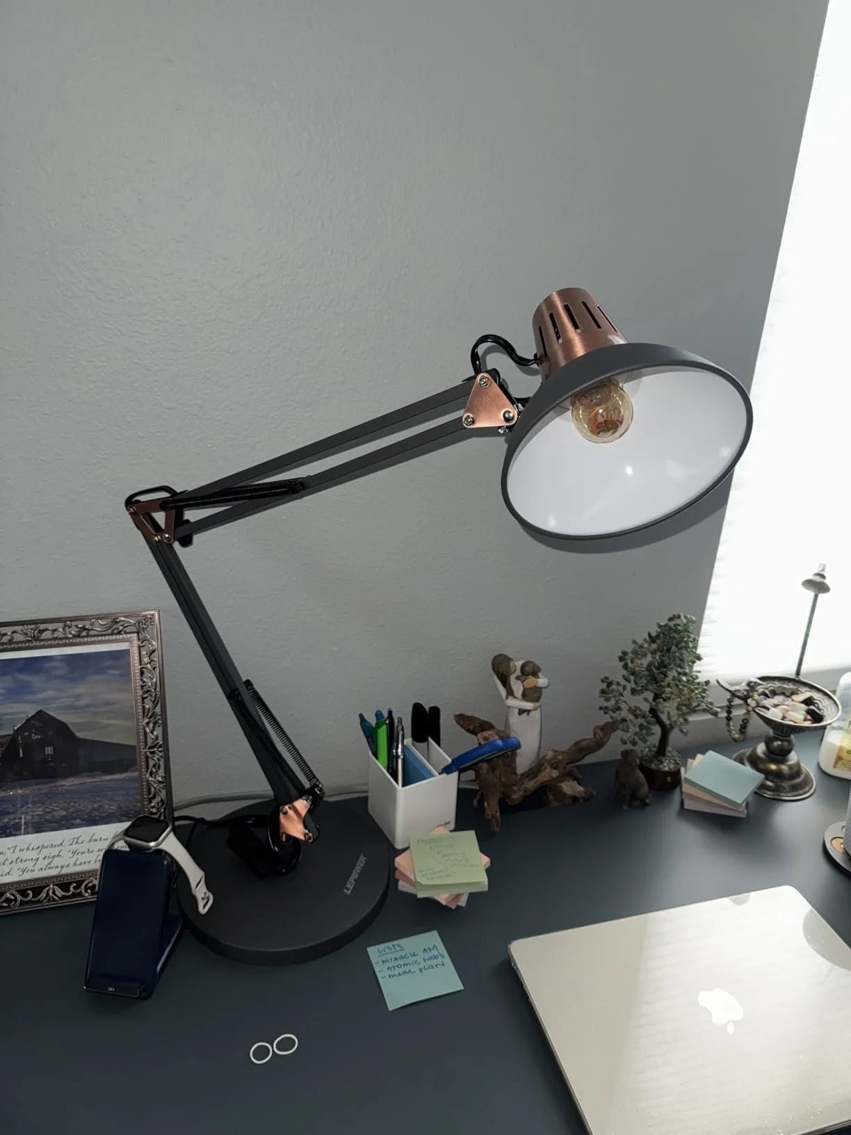 A black LEPOWER 16W LED Architect Lamp with a long, multi-jointed swing arm and a flat, rectangular LED panel, positioned on a dark desk with various office supplies and a laptop. The lamp has copper-colored accents.