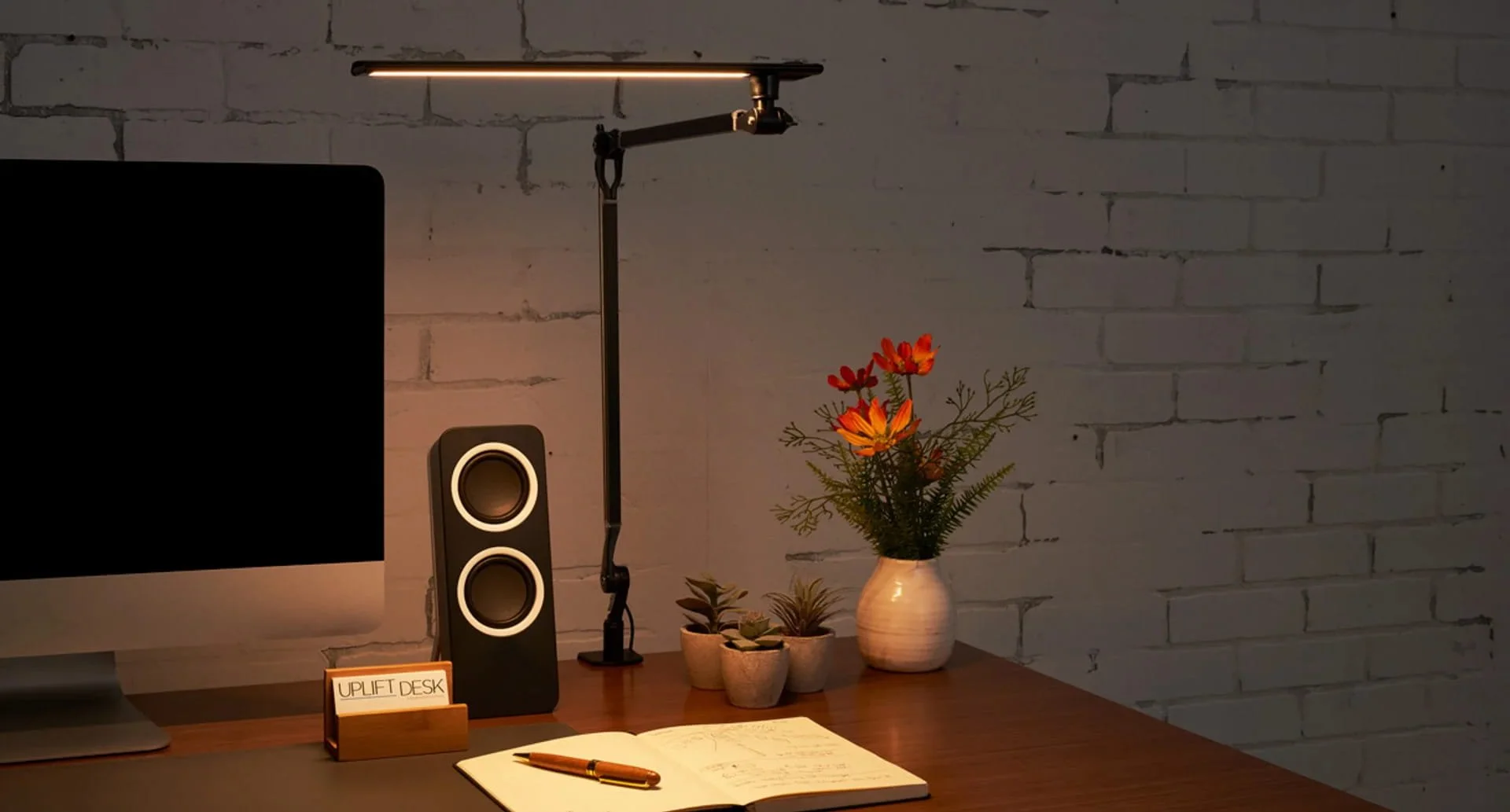 A black Uplift Desk E7 LED Clamp Lamp with an adjustable arm and horizontal light bar illuminates a desk with a computer monitor, speakers, a notebook, and a vase of flowers.