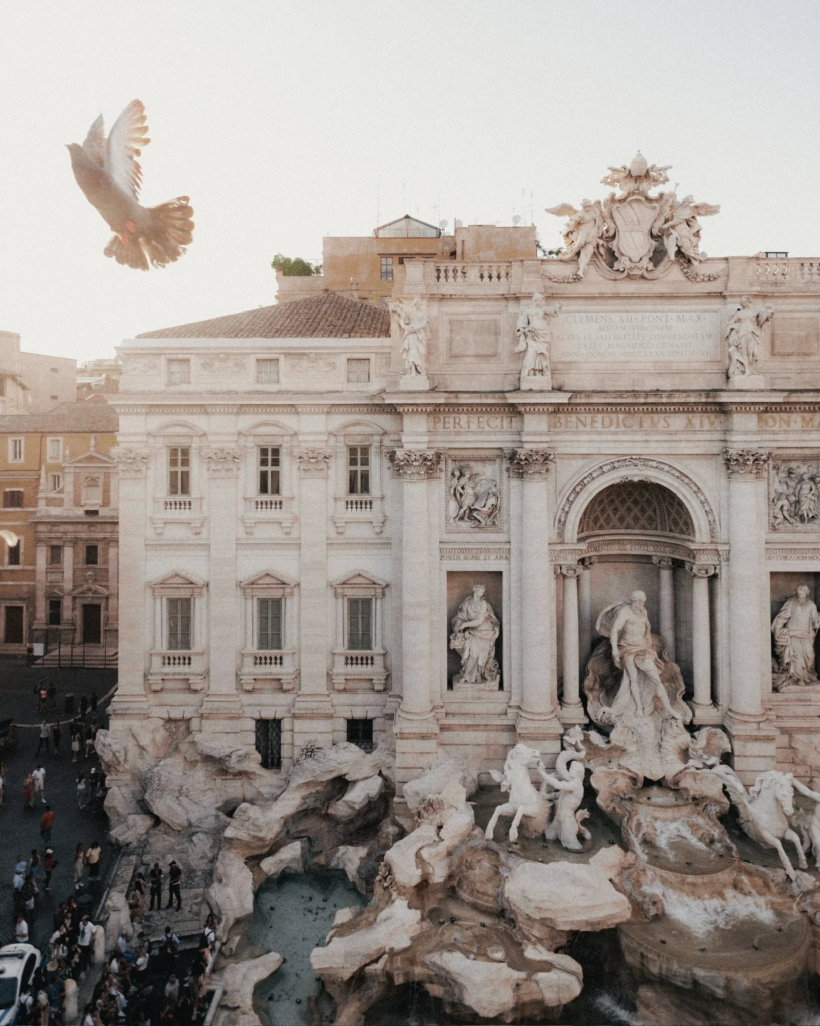 A historic fountain with sculptures and a building in the background, with a dove flying in the sky.