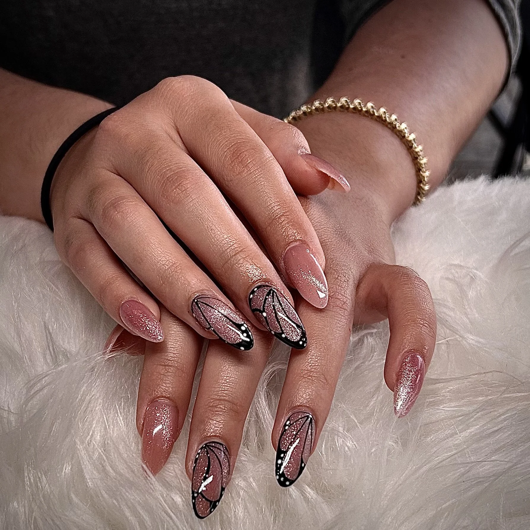 Close-up of hands with manicured nails, featuring pink glittery polish and black outlined abstract designs on some nails, resting on a white furry surface.