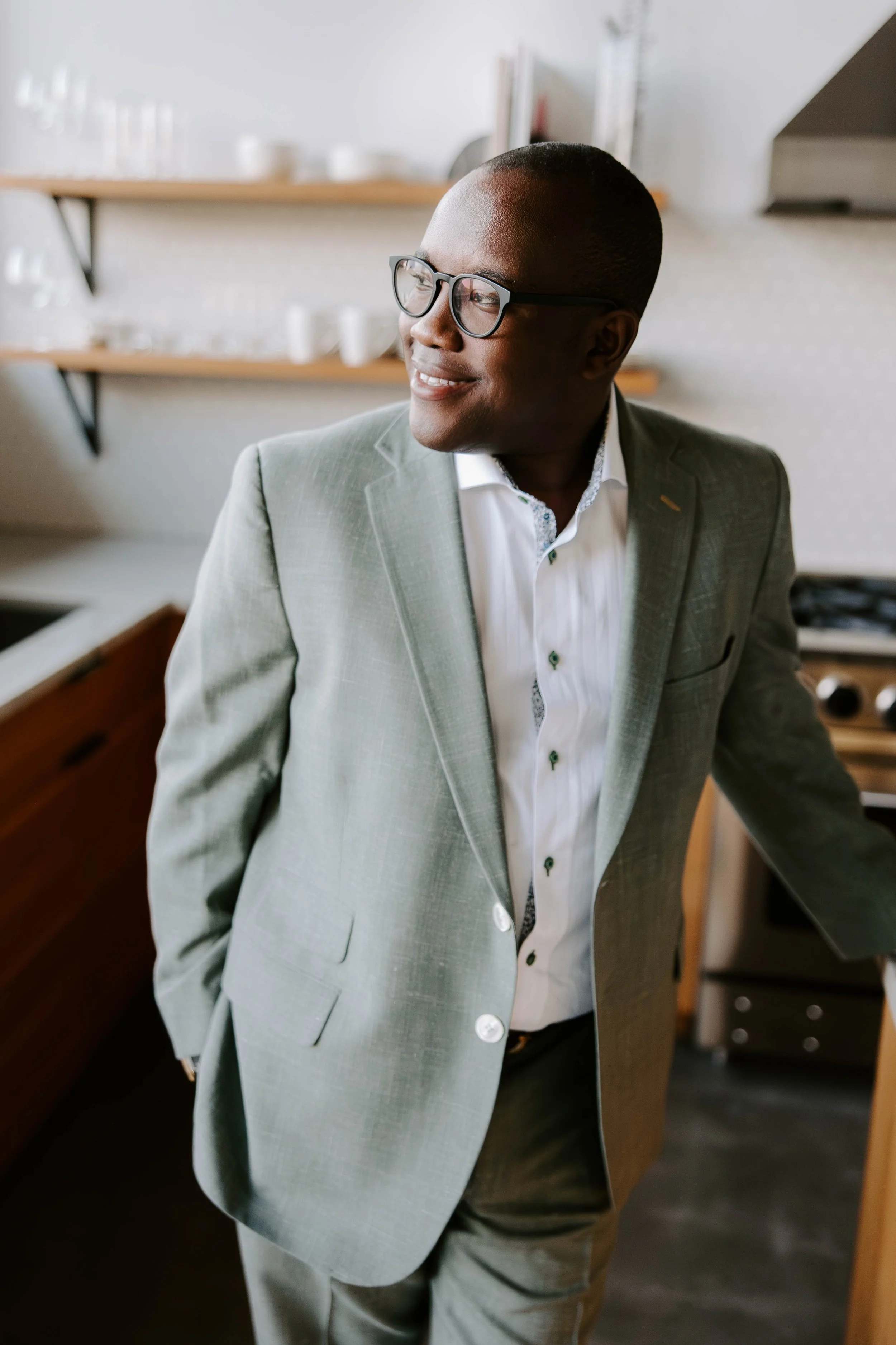 A man in a light gray suit and white shirt with dark glasses standing in a kitchen, looking off to the side and smiling.