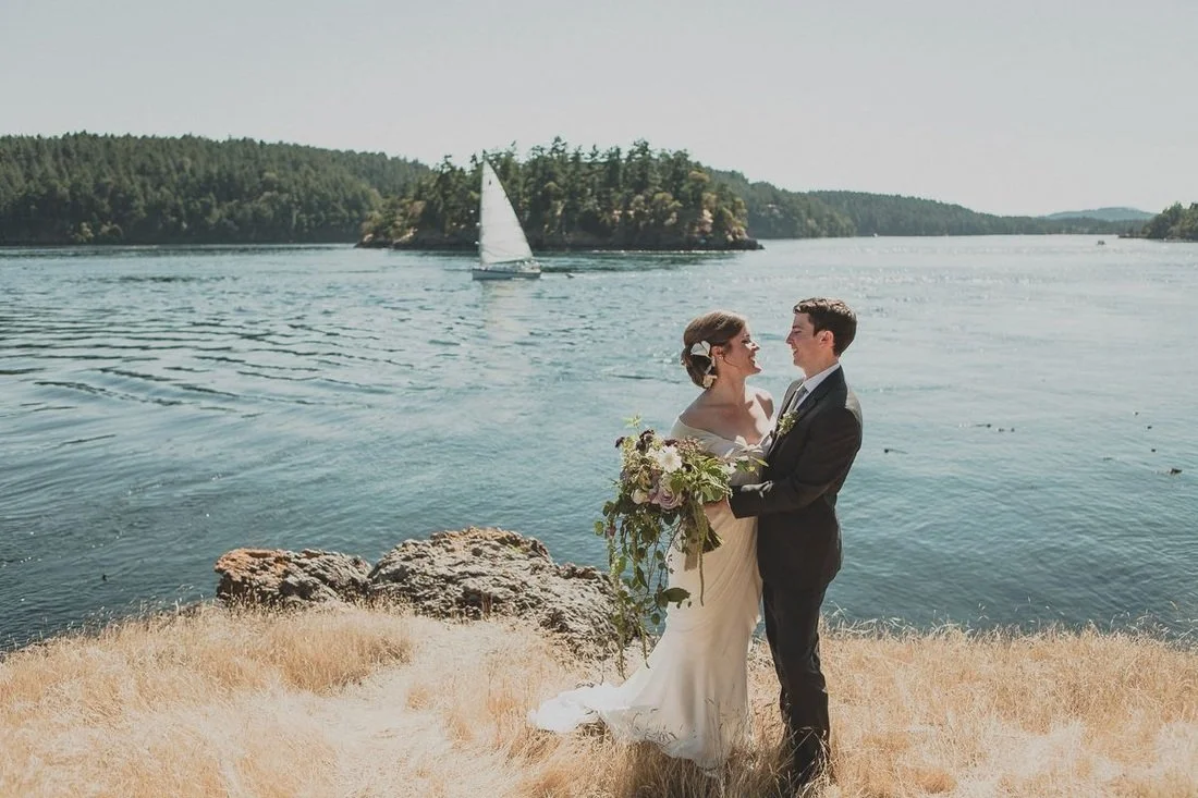 A bride and groom standing by a body of water, facing each other, with the bride holding a bouquet of flowers and the groom in a black suit. A sailboat is visible on the water in the background. Orcas island wedding, waterfront, westsound.